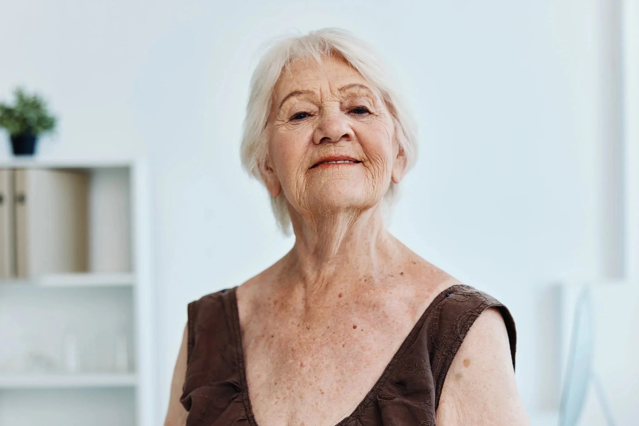 Ältere Frau mit weißen Haaren in einem dunklen Kleid steht in einem hellen Raum.