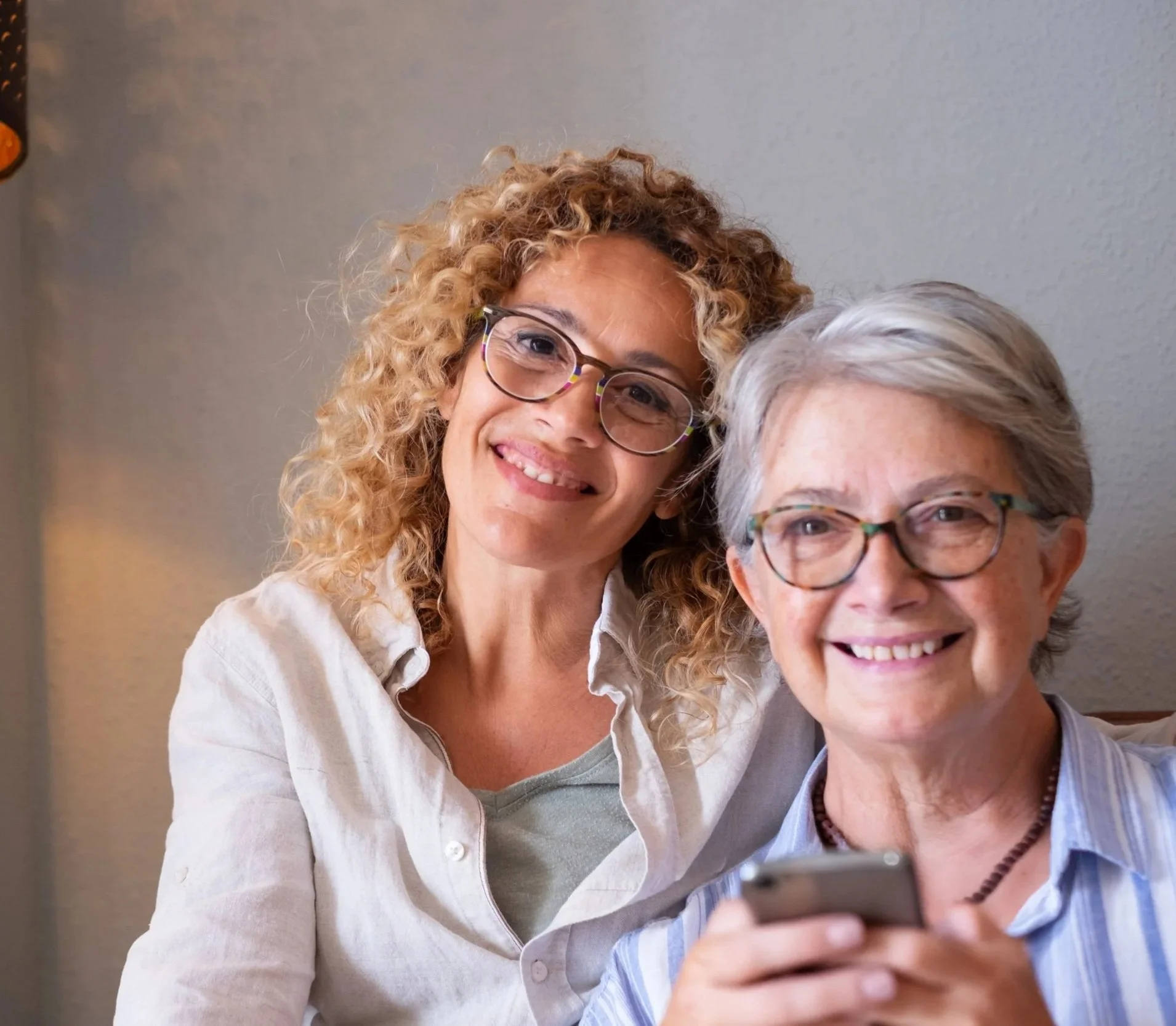 Zwei lachende Frauen, eine mit braunen lockigen Haaren und die andere mit kurzen grauen Haaren, posieren in einem Raum, eine hält ein Smartphone.