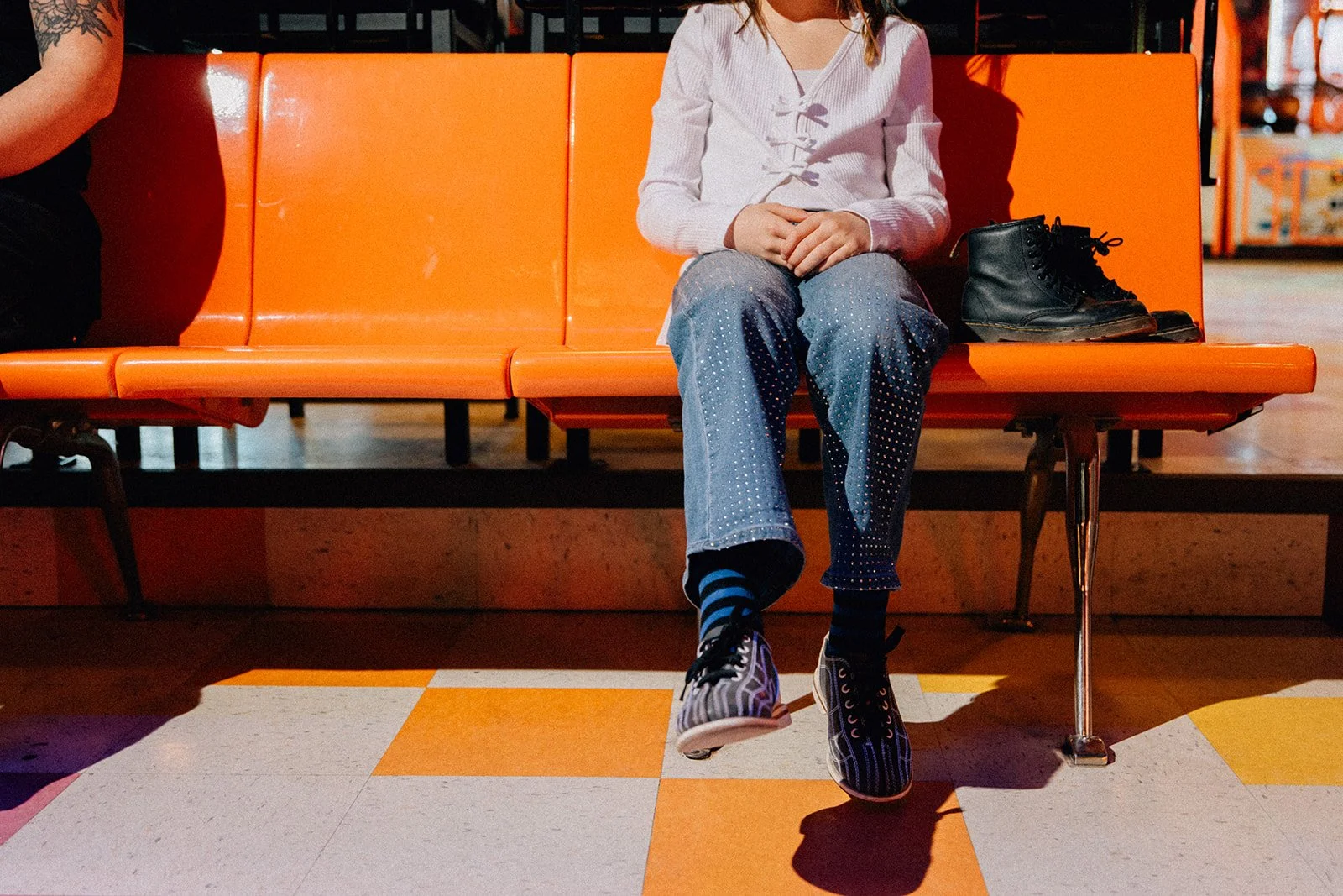 A person sitting on an orange bench in a public space with their legs crossed, wearing black and blue striped socks, patterned sneakers, and a purple hoodie. A pair of black boots is placed next to them on the bench.