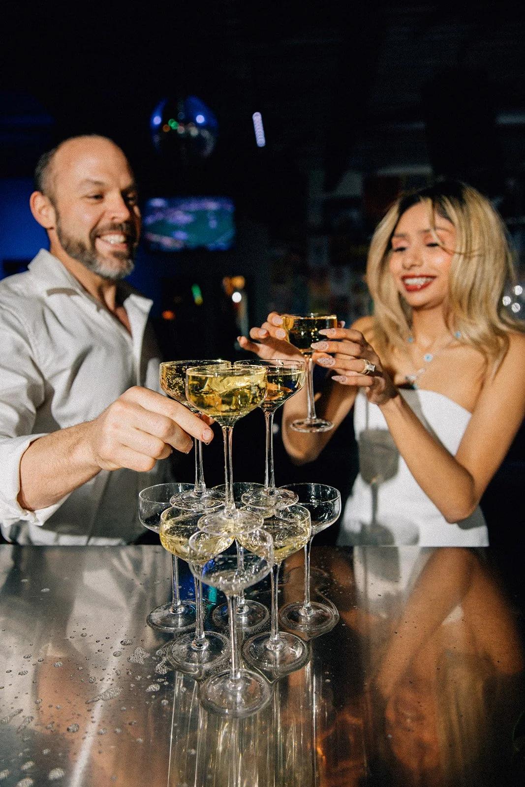 Man and woman clinking champagne glasses in a bar with a tower of stacked glasses on the counter.
