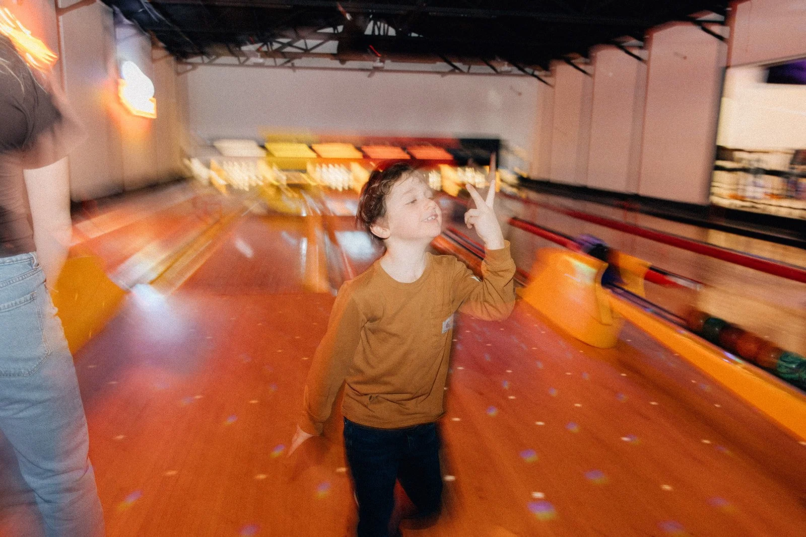 A boy at a bowling alley making a peace sign with his fingers, smiling, while an adult stands nearby. The background shows the bowling lanes, pins, and glow-in-the-dark effects.