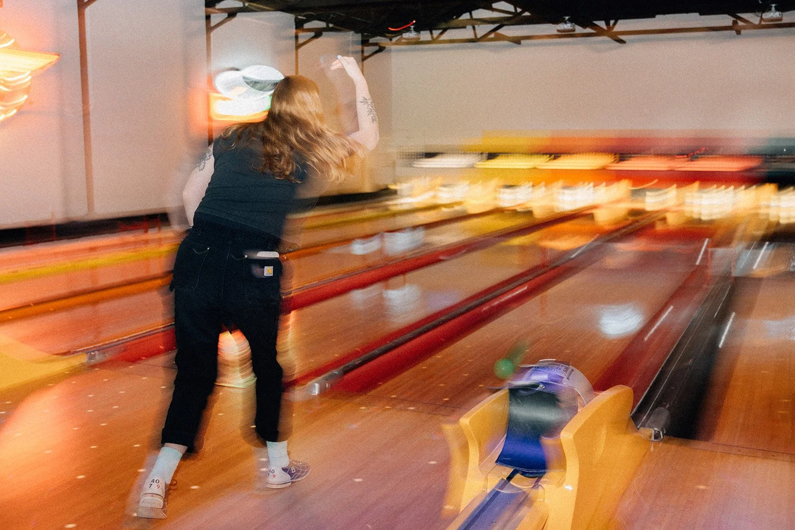 A woman with long hair, wearing a black shirt, black pants, white socks, and sneakers, is bowling at a bowling alley, with motion blur indicating movement.