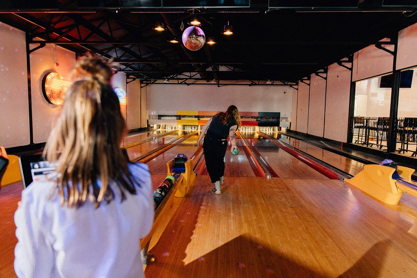 Two women are at a bowling alley, one is approaching the lane holding a bowling ball and the other is standing at the foreground watching, with her back to the camera.
