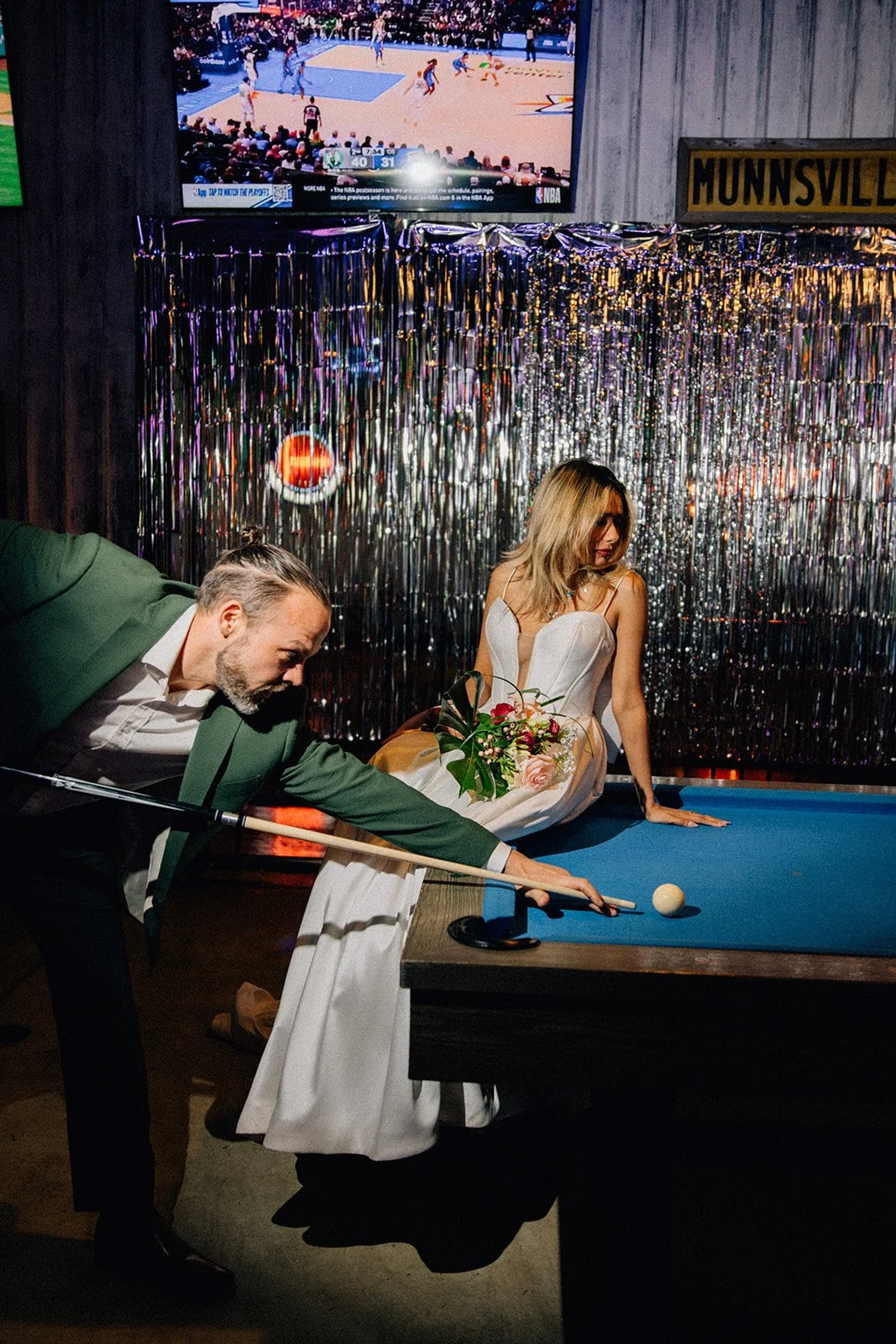 A man and woman playing pool in a decorated bar, with the woman sitting on the pool table holding a bouquet of flowers and the man taking his shot. A TV screen shows a basketball game in the background.