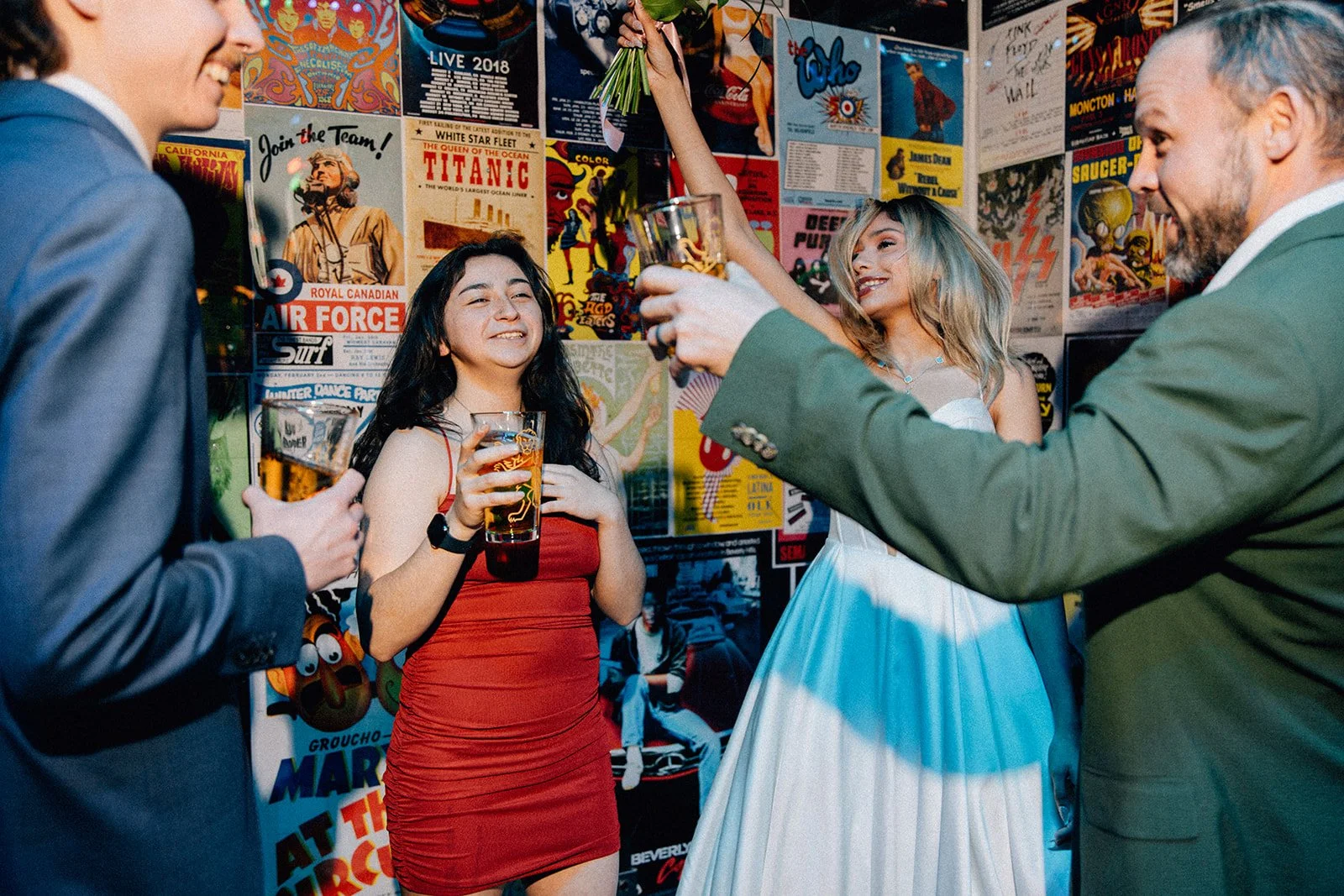Group of four people celebrating at a party with colorful posters on the wall behind them, two men and two women holding drinks, one woman wearing a red dress, the other a white dress.