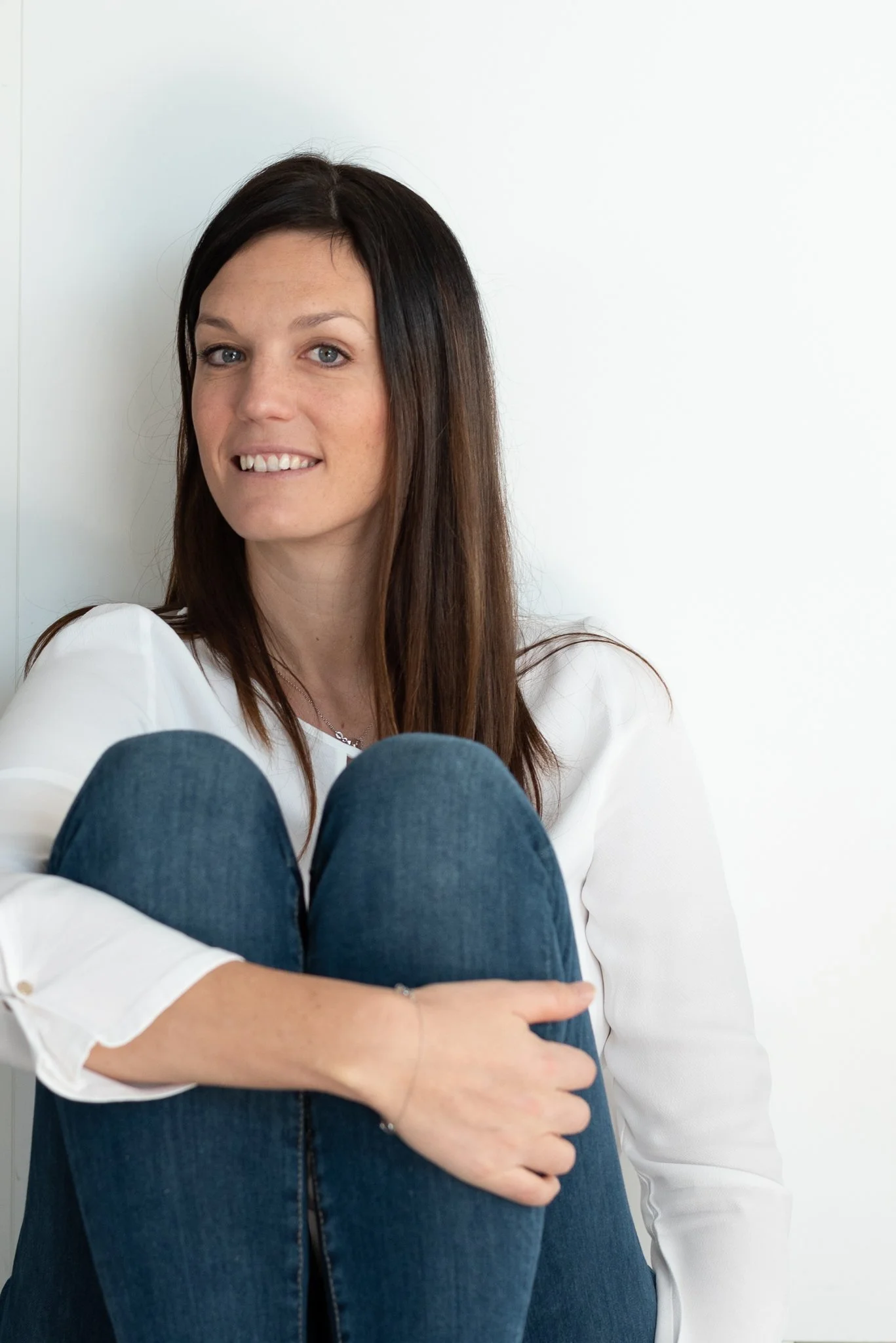 Jeune femme aux cheveux bruns, portant une chemise blanche, assise contre un mur blanc, souriant légèrement.