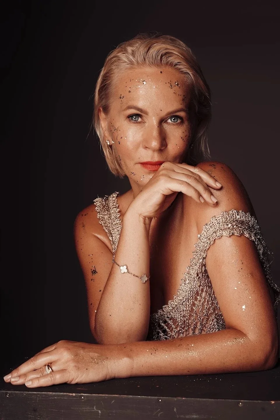 A woman with blonde hair, blue eyes, and light skin, adorned with black and silver glitter, is posing with her hand on her chin. She is wearing a sparkly dress and jewelry, with a dark background.