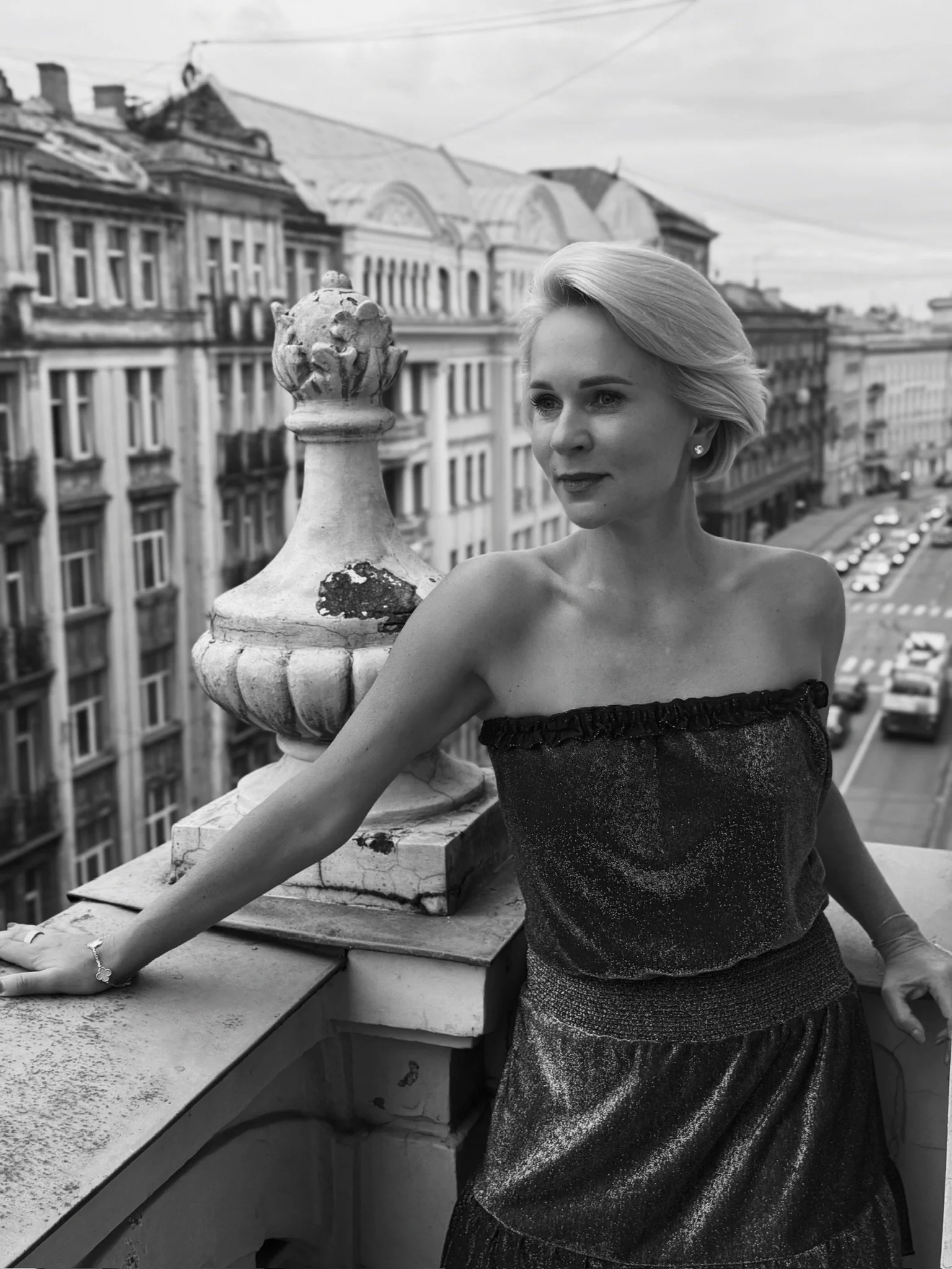 A woman with short blonde hair and a strapless dress standing on a rooftop balcony overlooking a city street, with old buildings and parked cars visible in the background, in black and white.