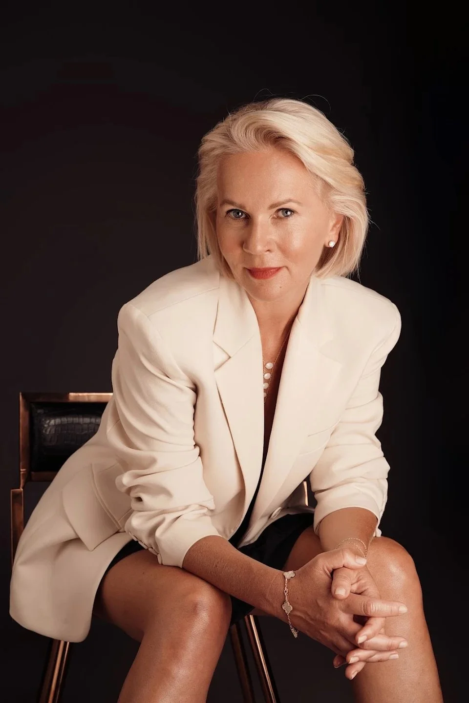 A woman with blonde hair in a cream blazer sitting on a chair against a black background.