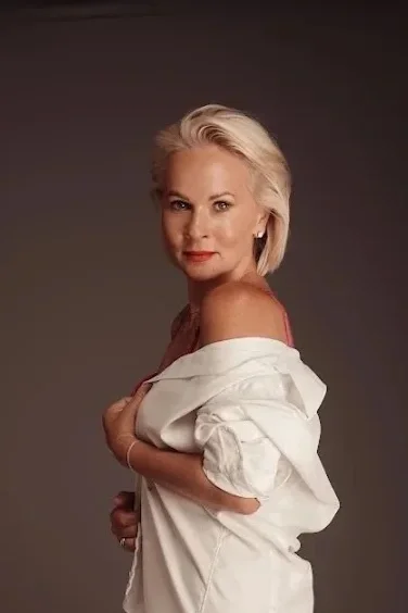A woman with short blonde hair wearing a white shirt slipping off her shoulder, set against a dark background.