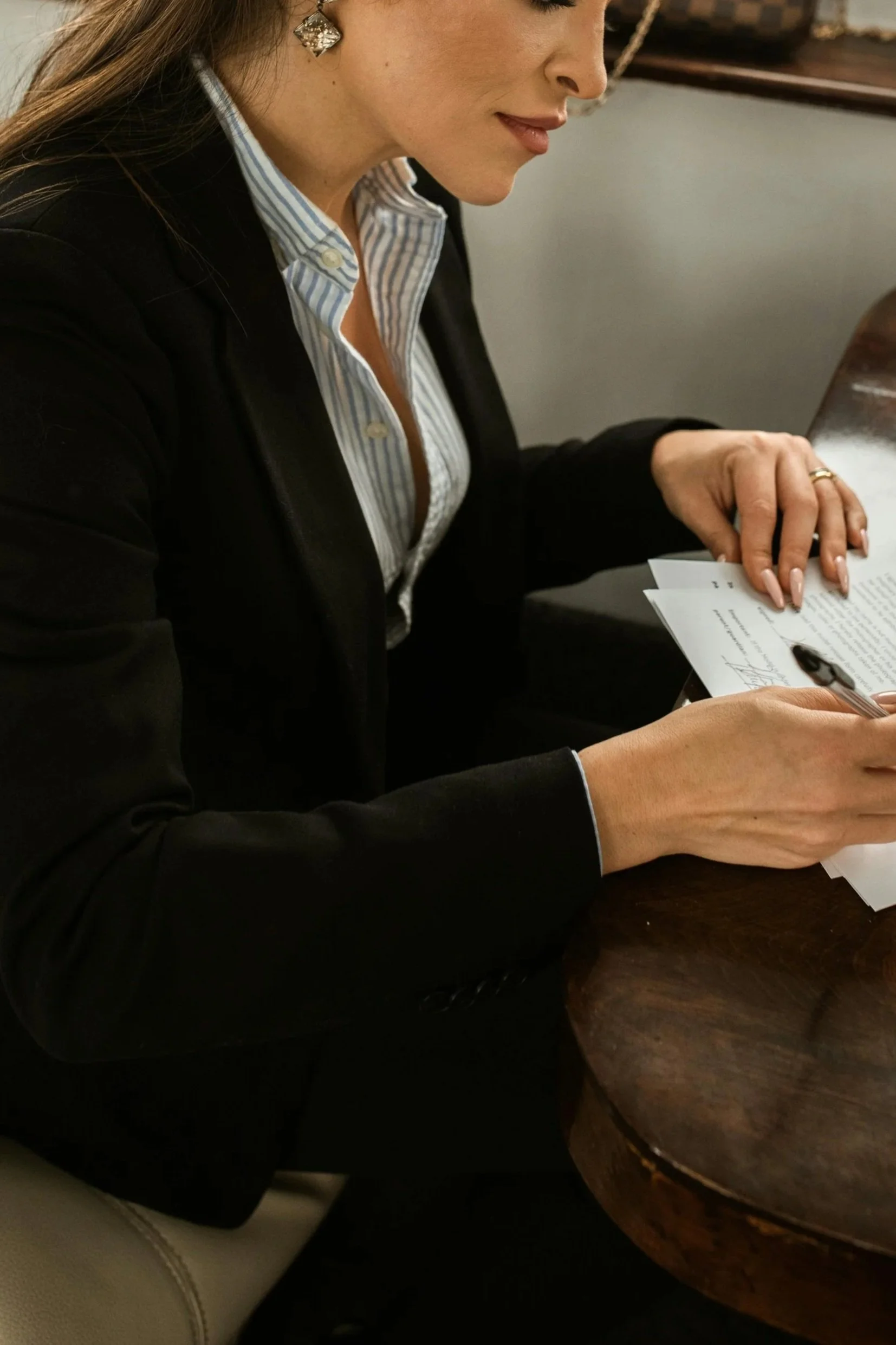 A woman in business attire sitting at a wooden table, signing documents with a black pen.