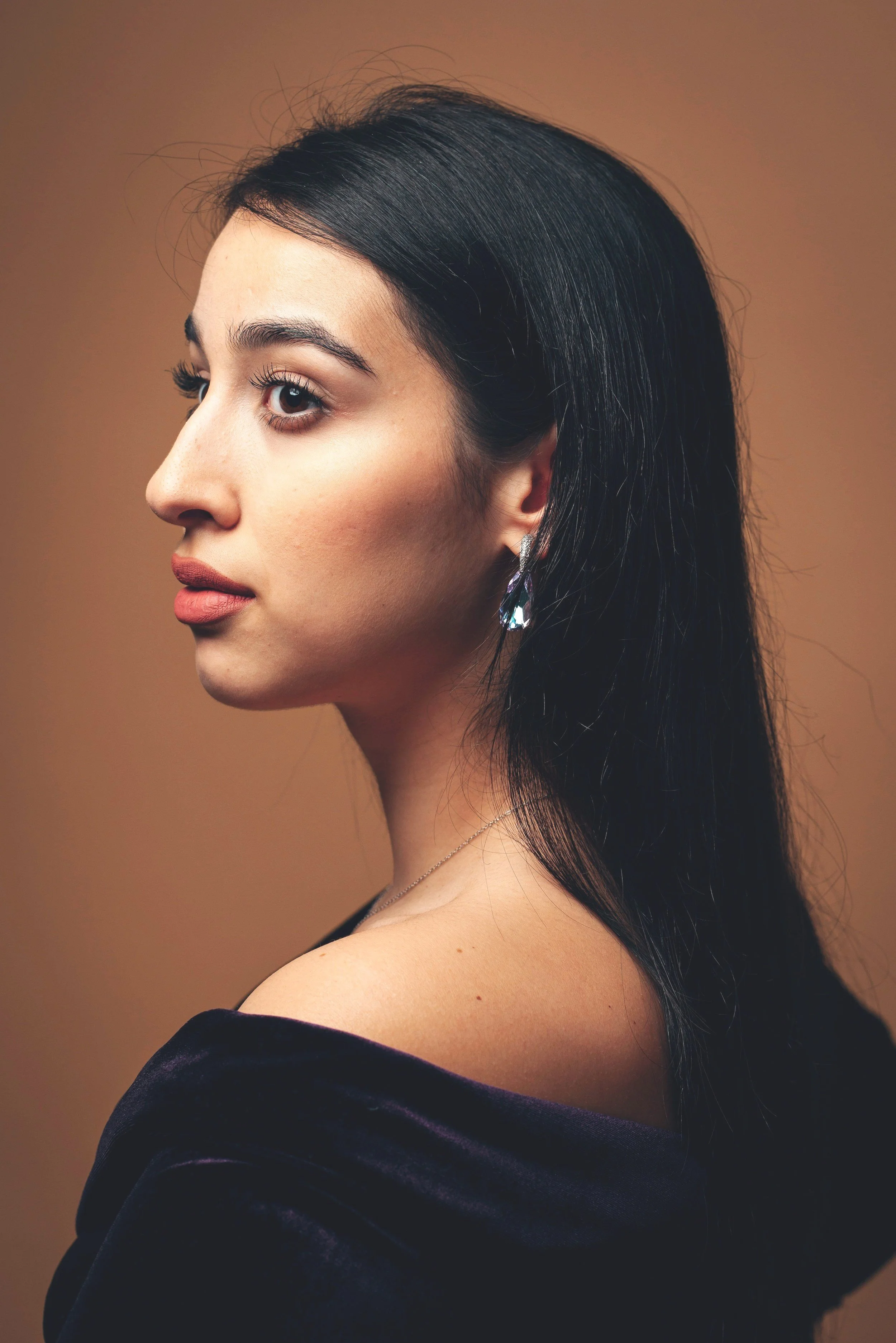 Side view of a woman with long dark hair, wearing earrings and a dark velvet top, against a plain brown background.
