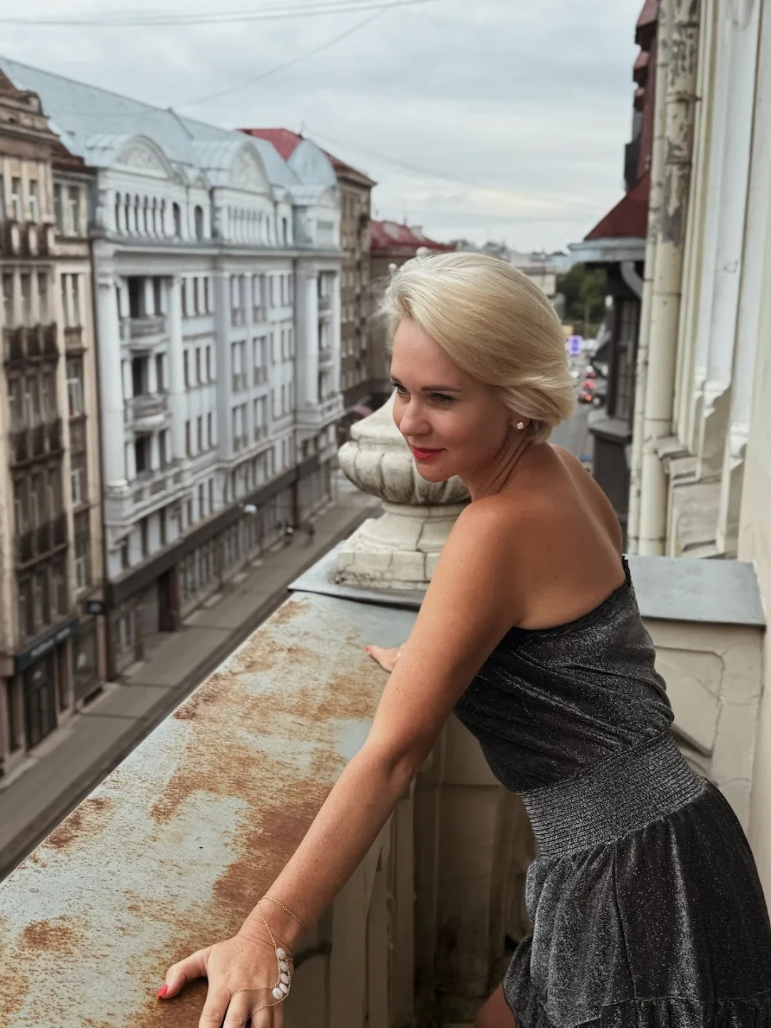 A woman with blonde hair wearing a strapless black dress leaning on a balcony railing, overlooking a city street with historic buildings.