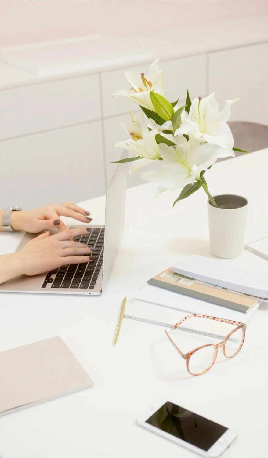 A person working on a laptop at a white desk with a vase of white lilies, glasses, notebooks, a gold pen, and a smartphone.