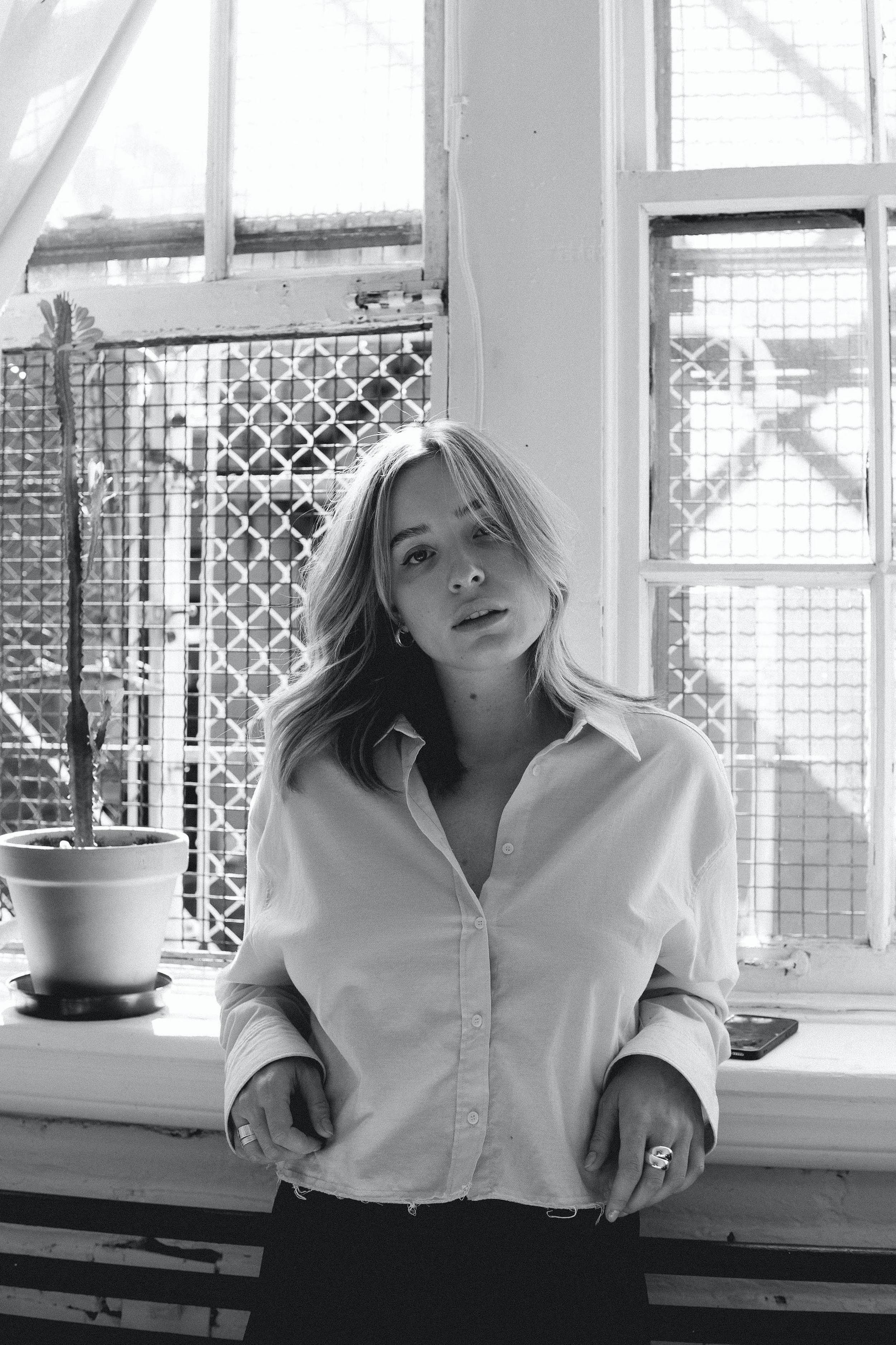 A woman with shoulder-length hair wearing a light-colored unbuttoned shirt standing in front of a large window with a grid pattern and a potted cactus plant on the windowsill.