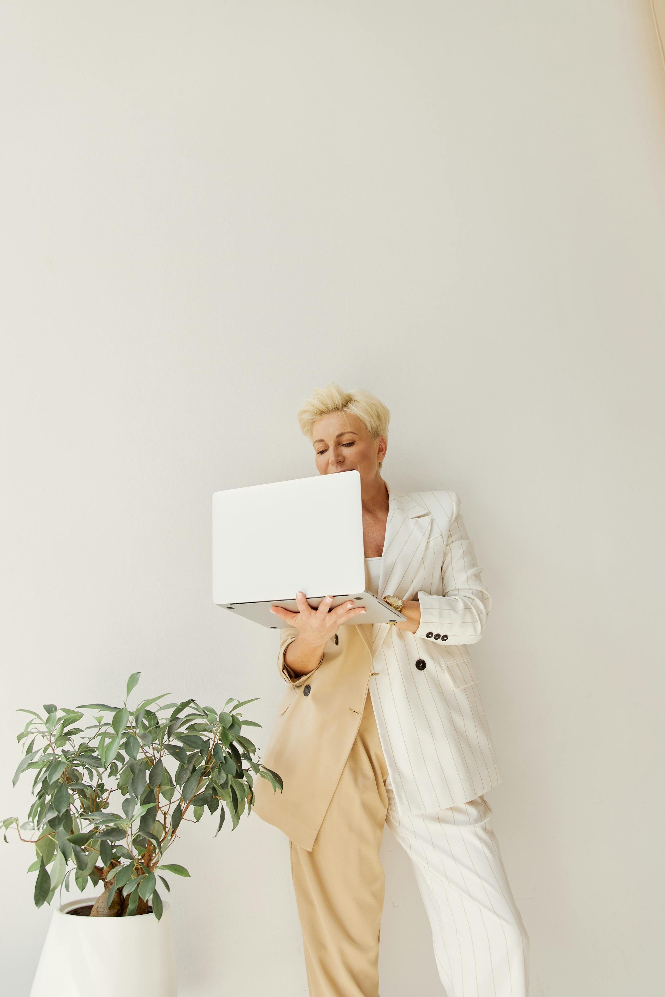 Woman in a cream and white pinstripe suit holding a laptop, standing next to a potted plant, against a plain light-colored wall.