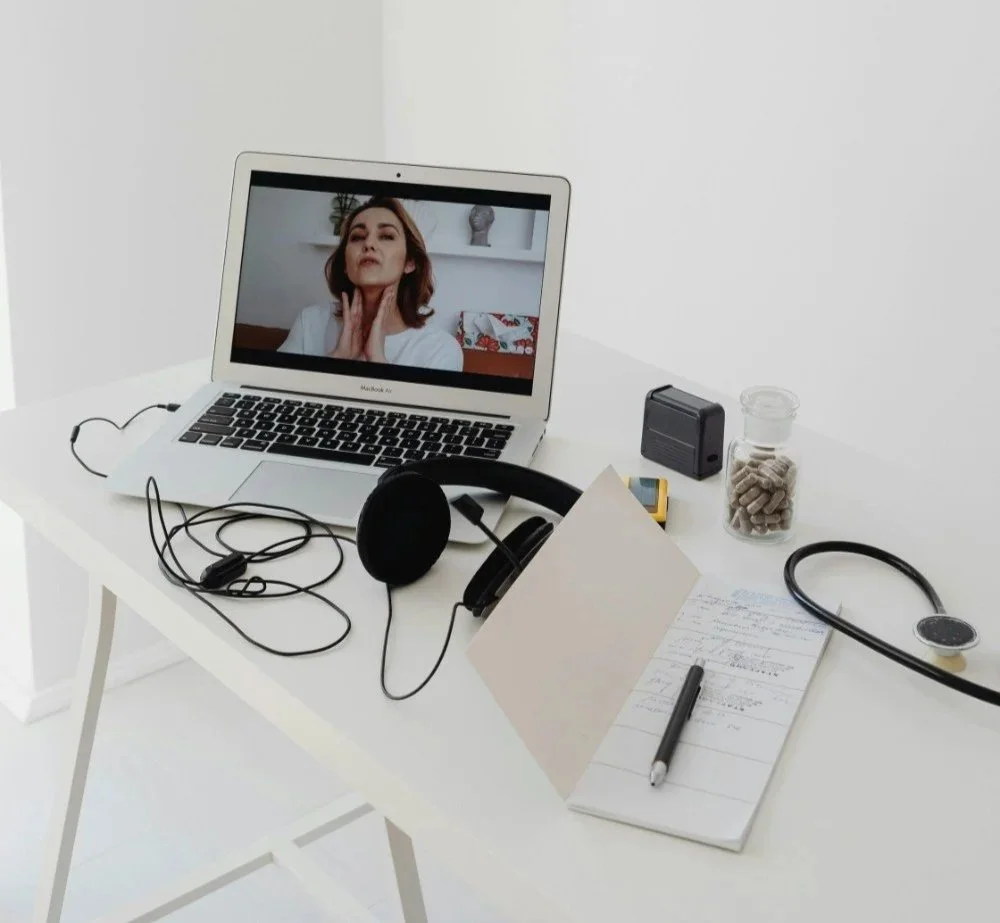 A white desk with a laptop showing a woman on a video call, a headset, a notebook with a pen, a stethoscope, a capsule bottle, a tape measure, a speaker, and a container of pills.