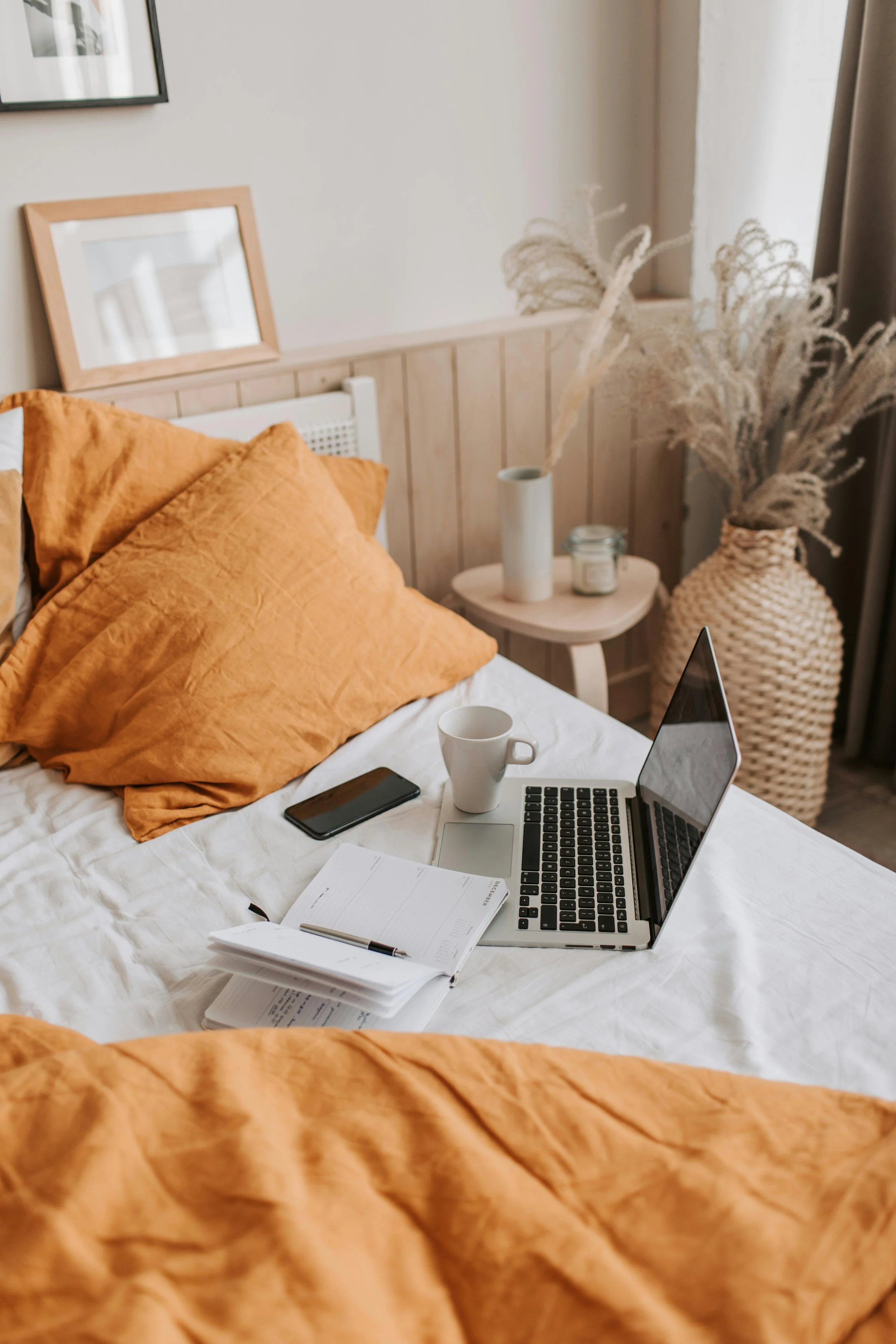 A neatly made bed with orange bedding, a laptop, a white mug, a smartphone, an open notebook, and a pen. A side table with vases and dried plants is in the background.