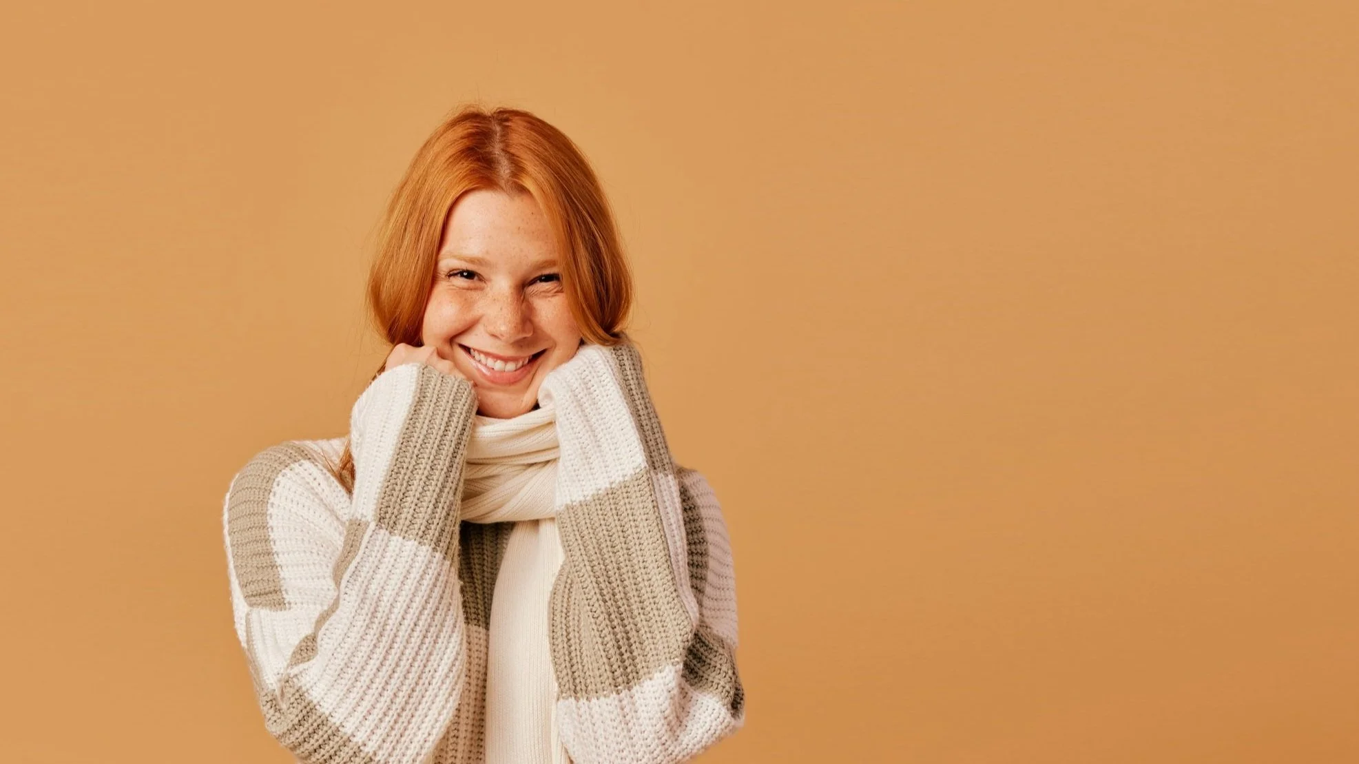 A young woman with red hair and freckles, smiling, wearing a white and beige striped sweater and a white scarf, standing against a plain beige background.