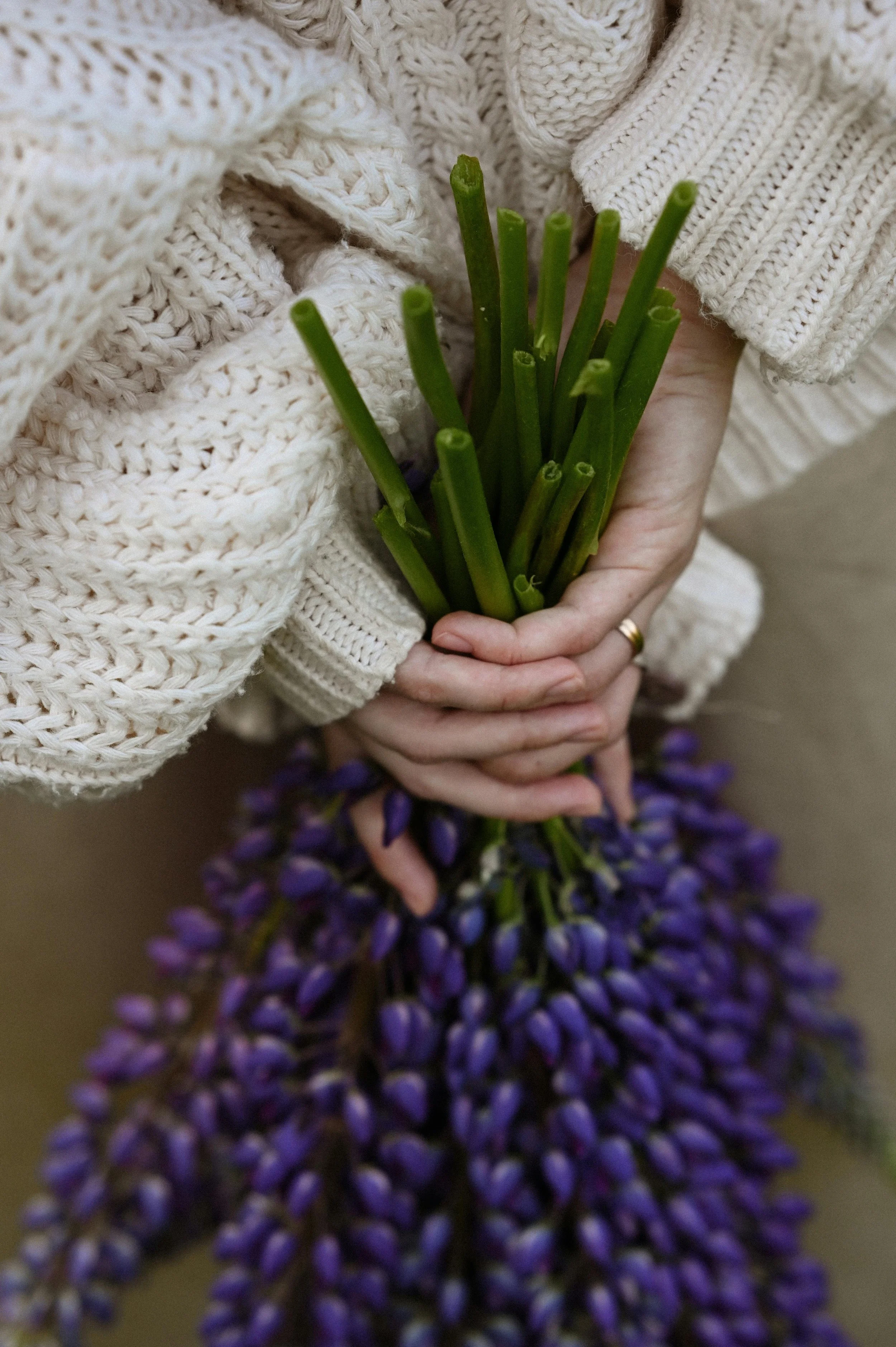 Person in a cream-colored knitted sweater holds a bunch of purple and green flowers.