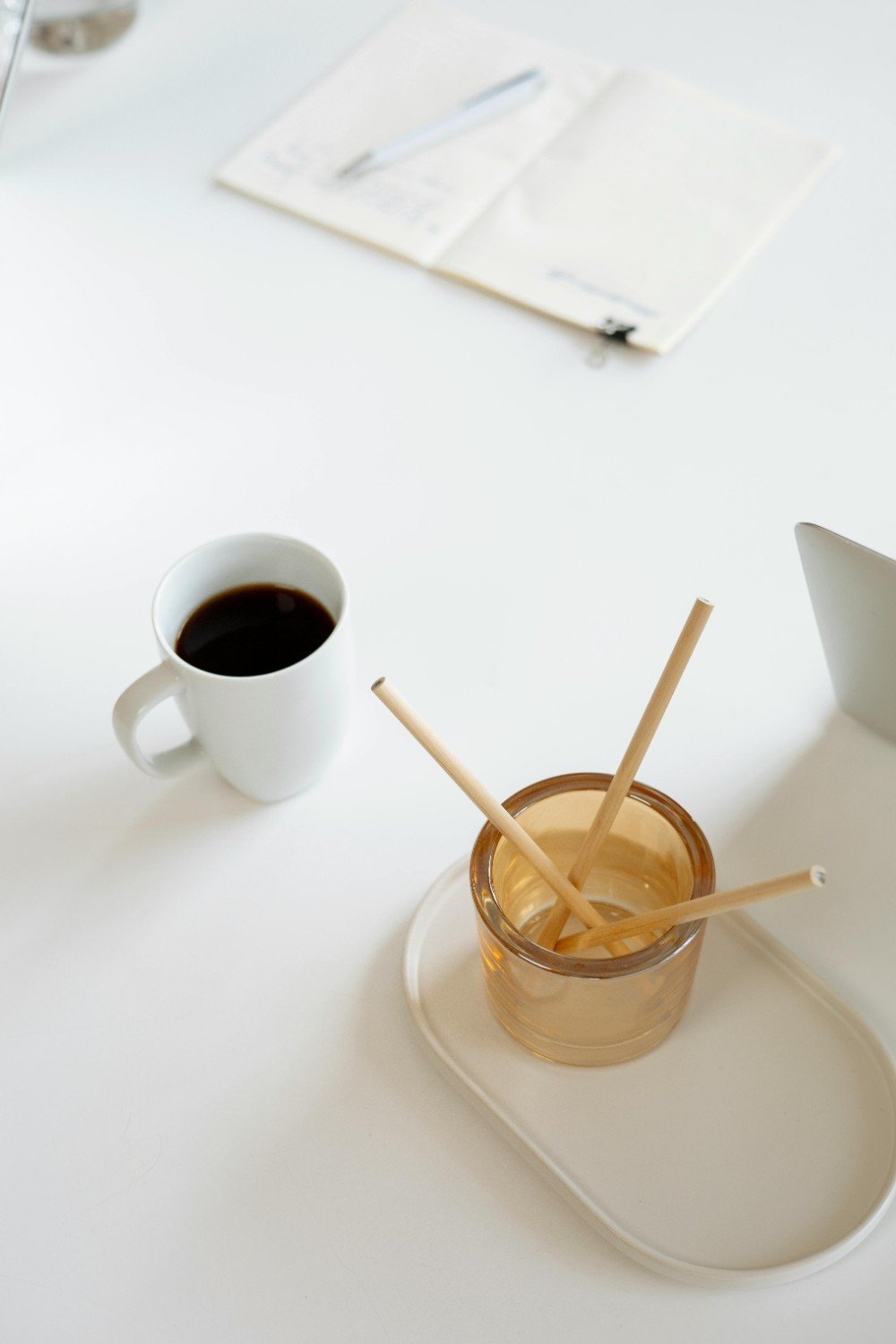A white table with a white coffee mug filled with black coffee, a glass container holding four wooden stir sticks, a white tray, a notebook with a pen, and a white pitcher or container.