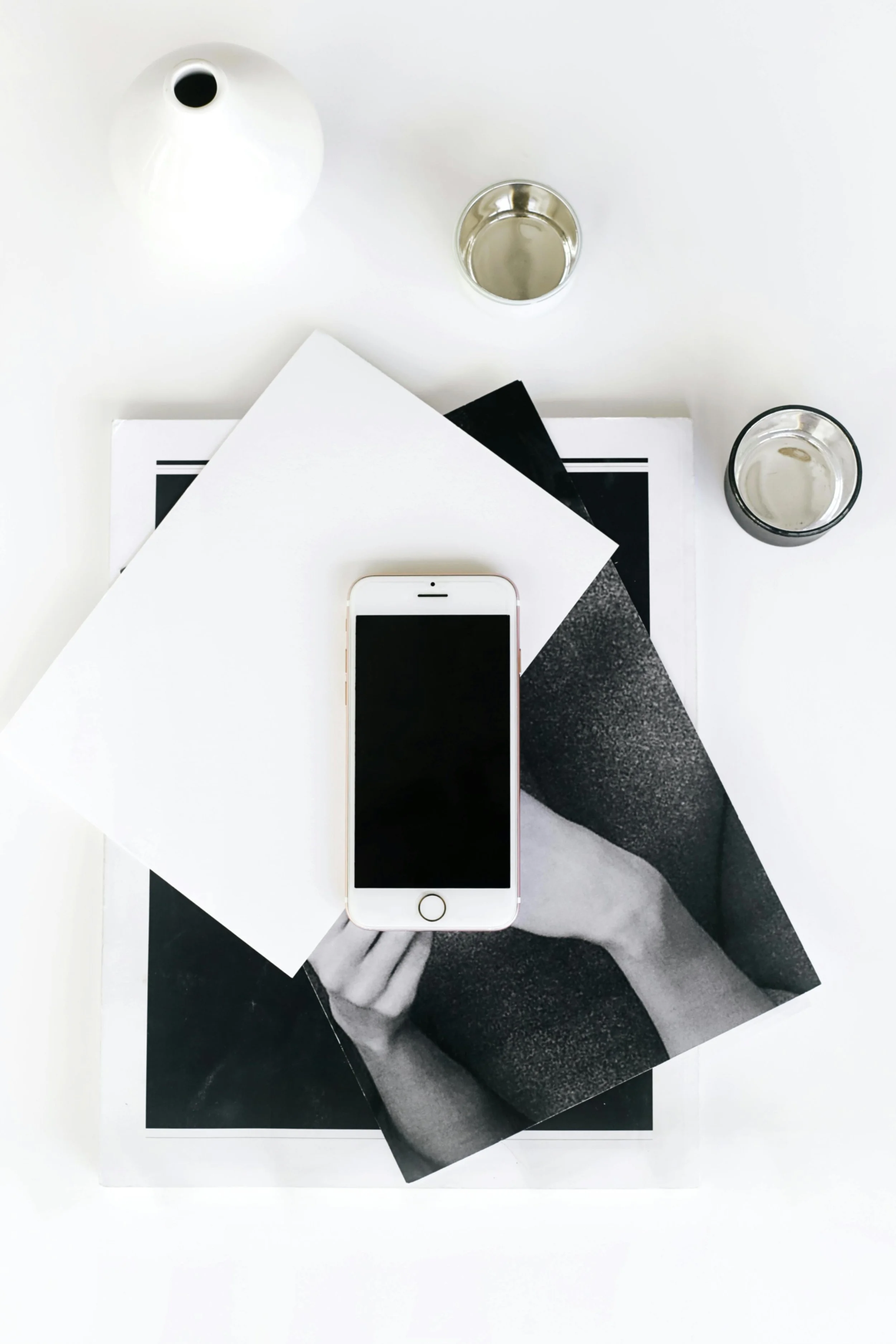 Flat lay of a white table with a white vase, two glasses of water, a smartphone, a black and white photo, and a magazine.