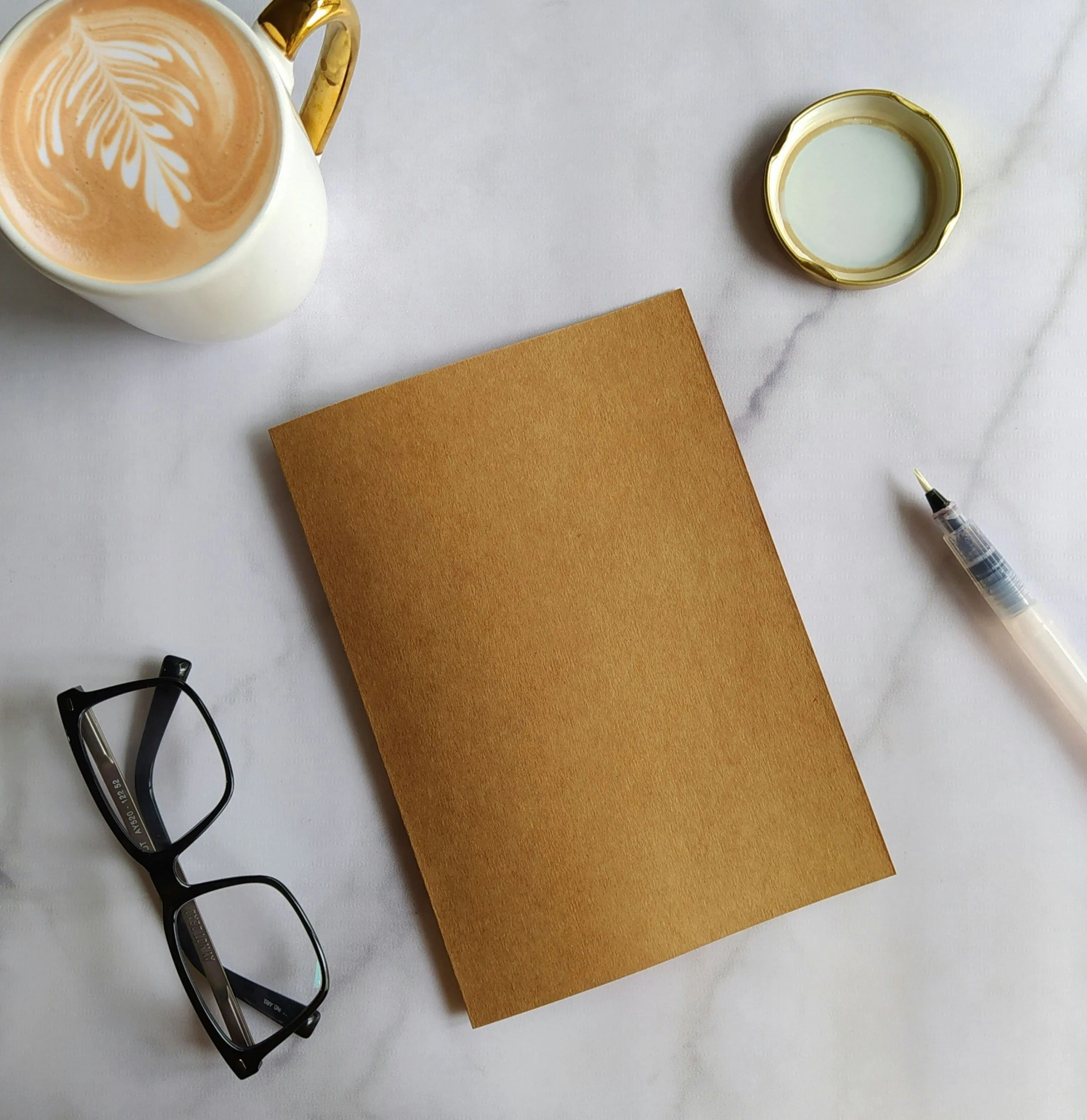 A flat lay of a white table with a notebook, a pair of glasses, a latte in a white and gold mug, a pen, a small dish, and a cup of cream or milk.