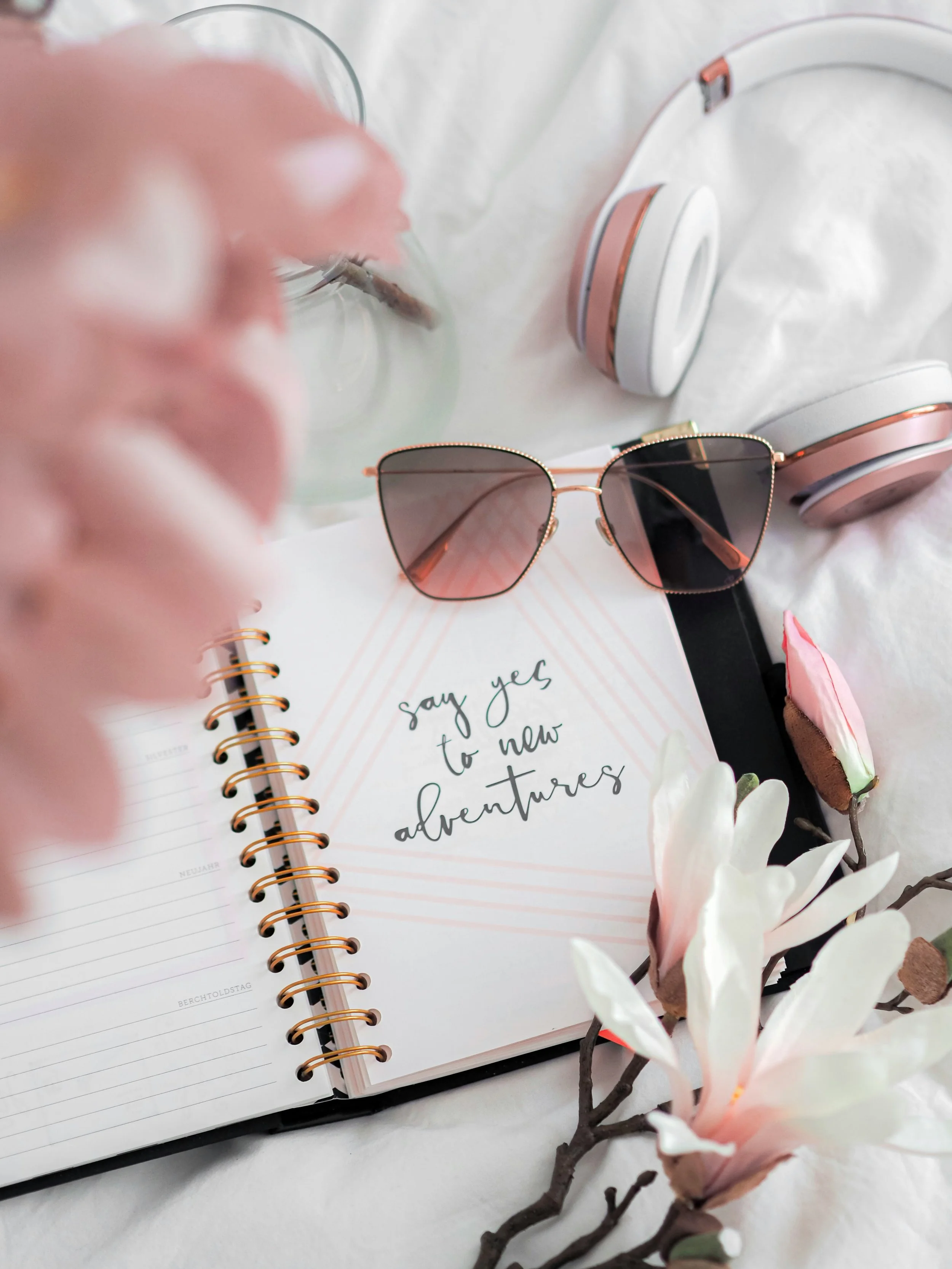 A journal with the quote "say yes to new adventures," pink magnolia flowers, pink and white headphones, a glass of water, and sunglasses on a white surface.