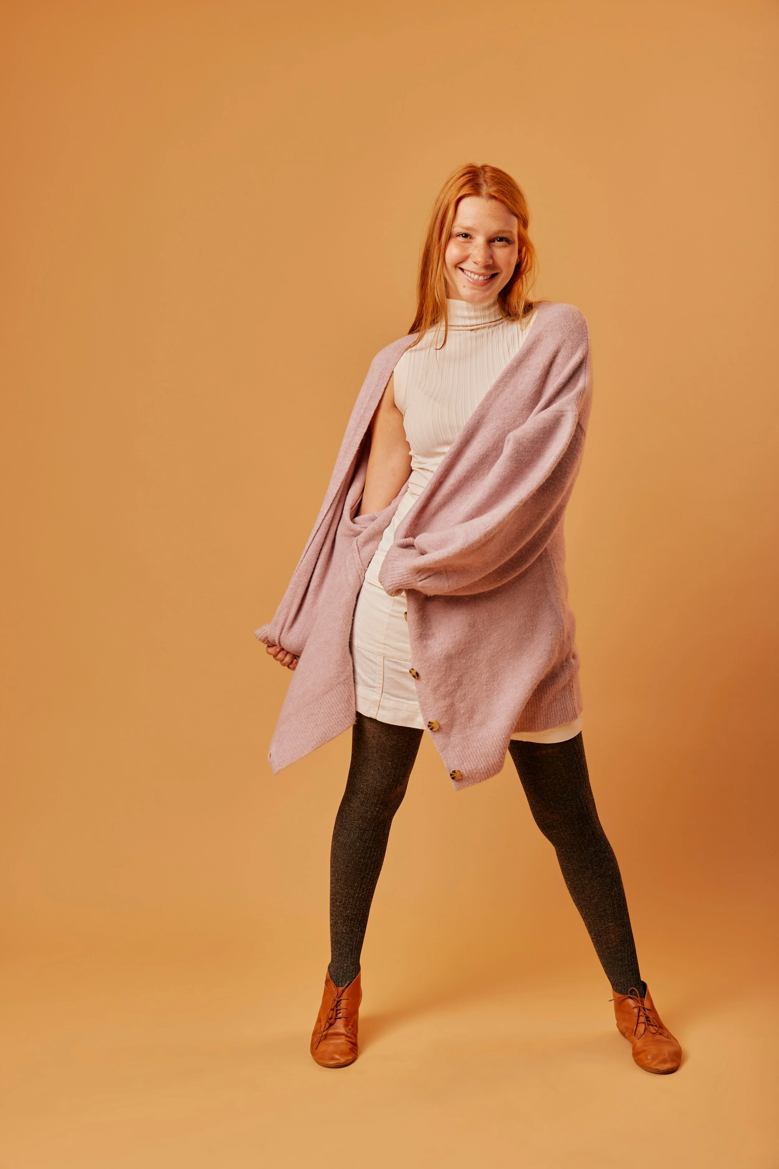 A woman standing with her legs apart, smiling, wearing a beige sleeveless turtleneck, pink oversized cardigan, beige skirt, black tights, and brown ankle boots, against a plain beige background.