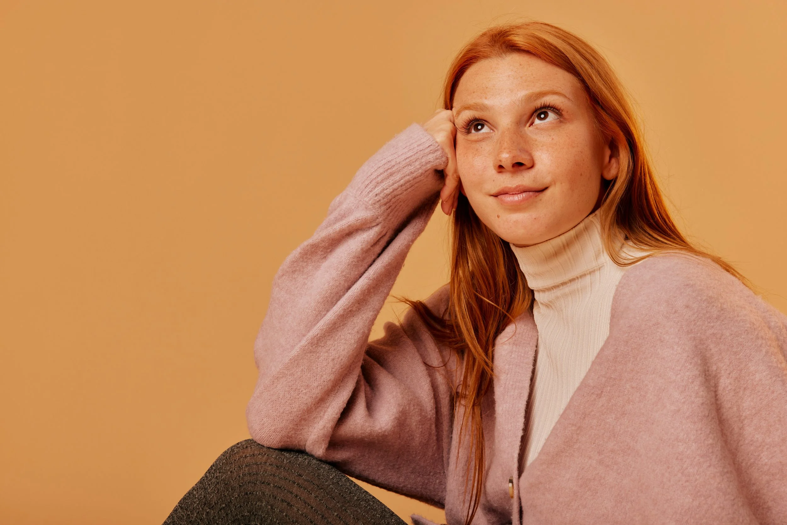 A young woman with red hair and freckles wearing a beige turtleneck sweater and pink coat, sitting against a beige background.