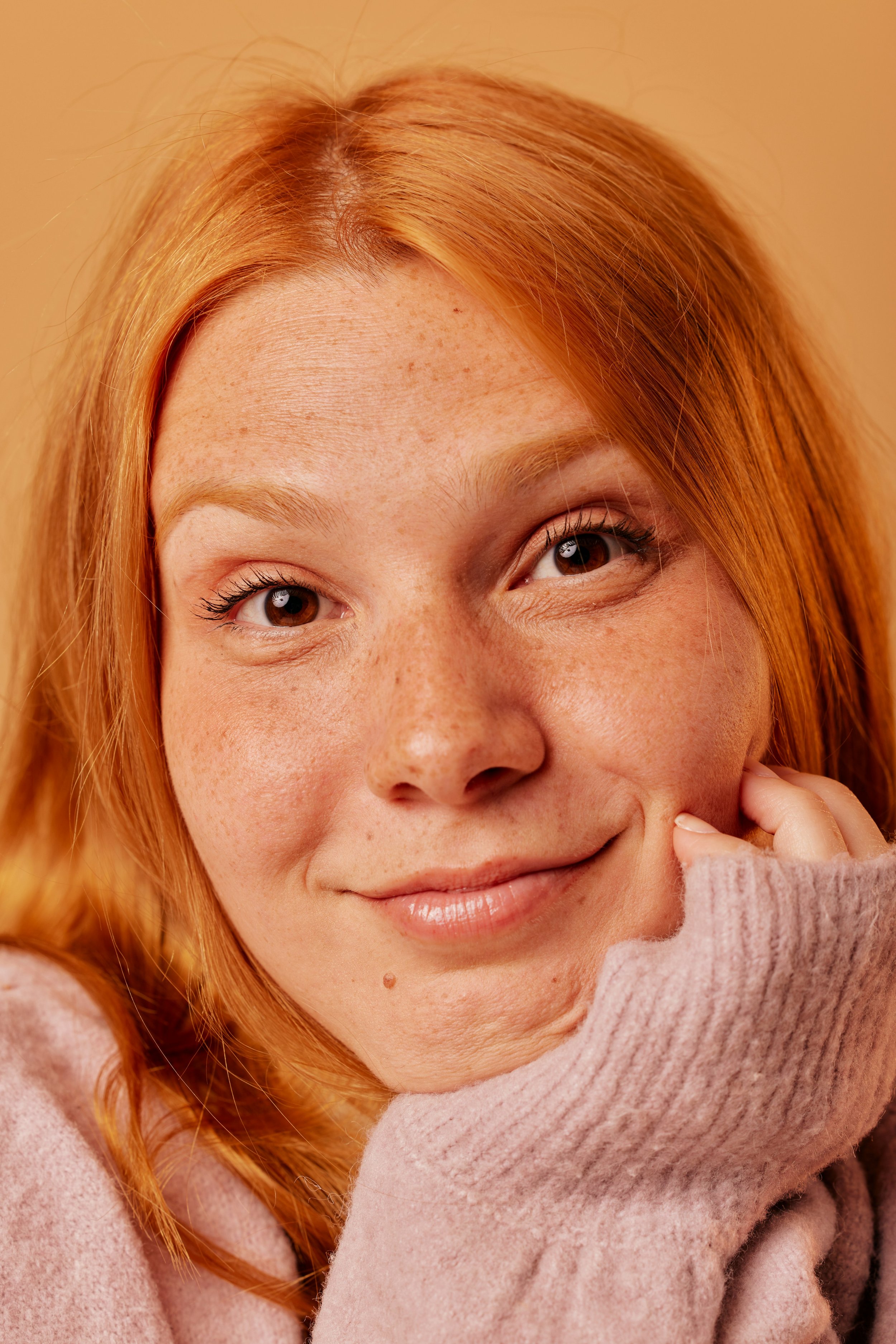 Close-up of a smiling woman with red hair and freckles, wearing a pink sweater, resting her chin on her hand.