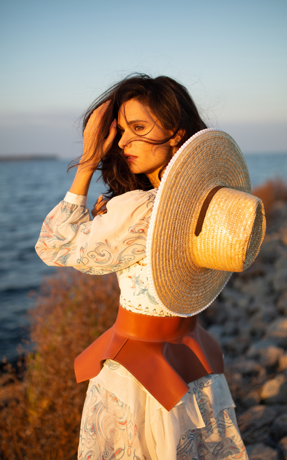 Woman by the water holding a wide-brimmed straw hat, wearing a cream-colored embroidered blouse and a brown leather belt skirt during sunset.