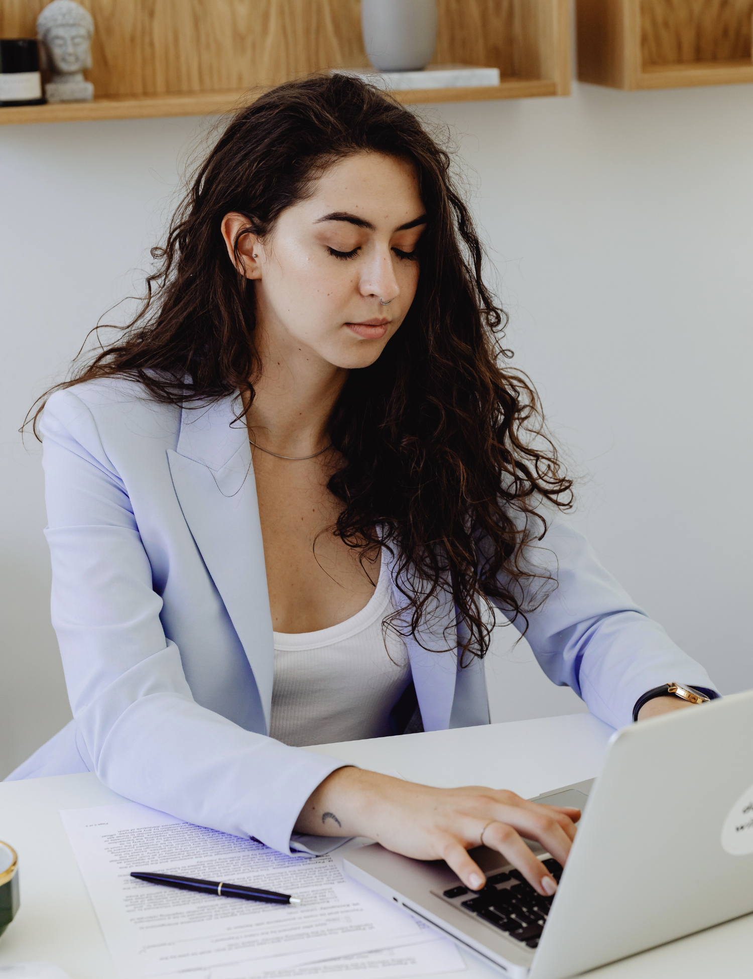 A woman with long curly brown hair, wearing a white tank top and light blazer, works on a laptop at a desk with documents and a pen.