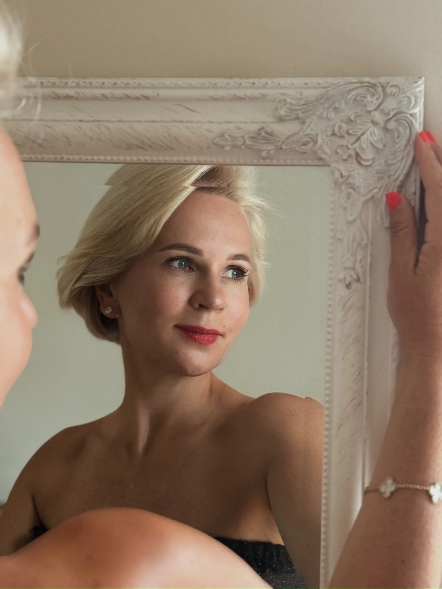 A woman with blonde hair and red lipstick looking into an ornate white mirror.
