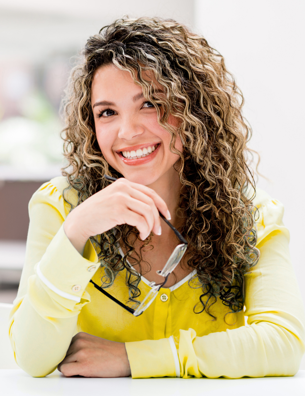 A woman with curly hair wearing a yellow top, smiling and holding reading glasses in her hand.