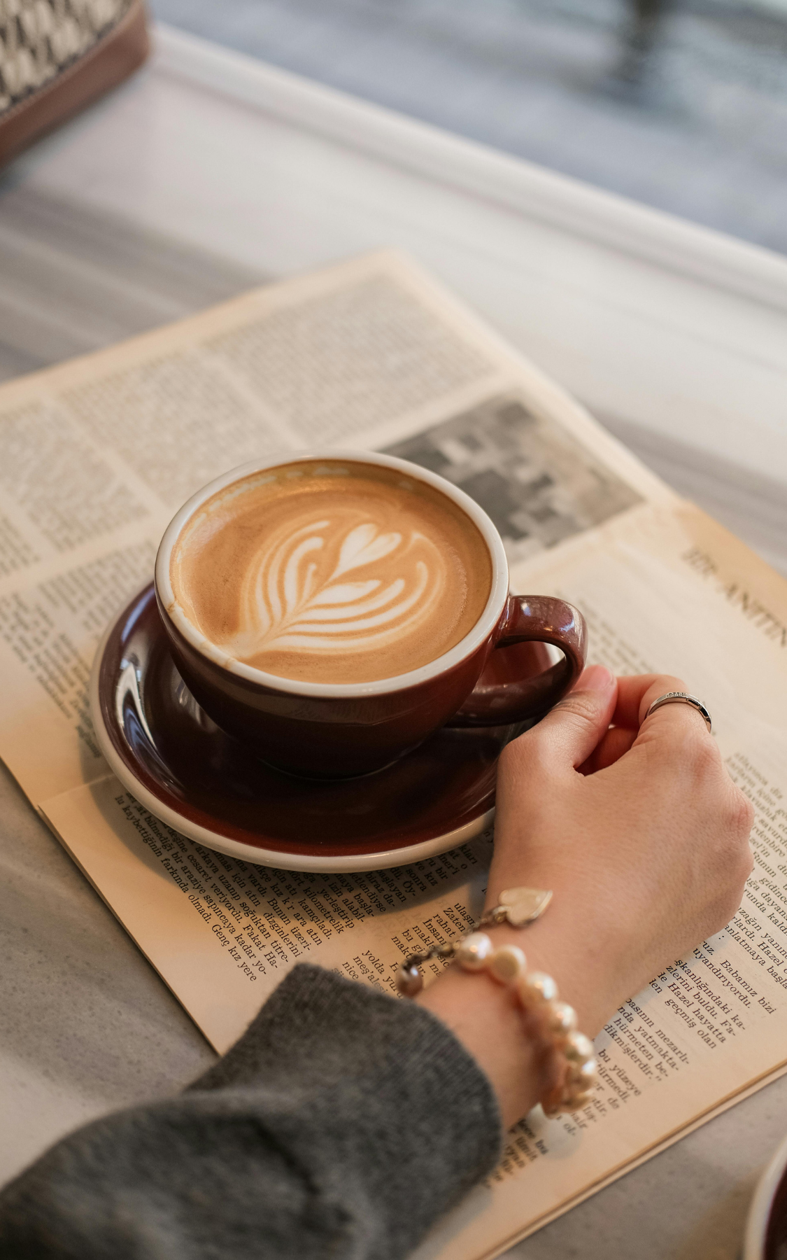 A cup of latte with latte art on top, placed on a saucer on a newspaper, with a person's hand touching the cup, wearing jewelry, on a table near a window.
