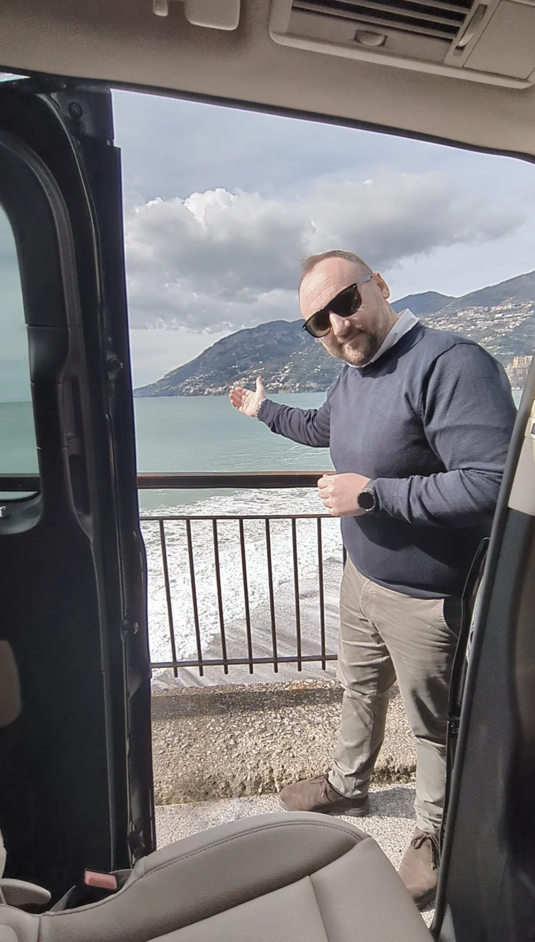 A man in sunglasses and a gray sweatshirt is standing outside a vehicle, pointing at a lake with mountains in the background, near a railing.
