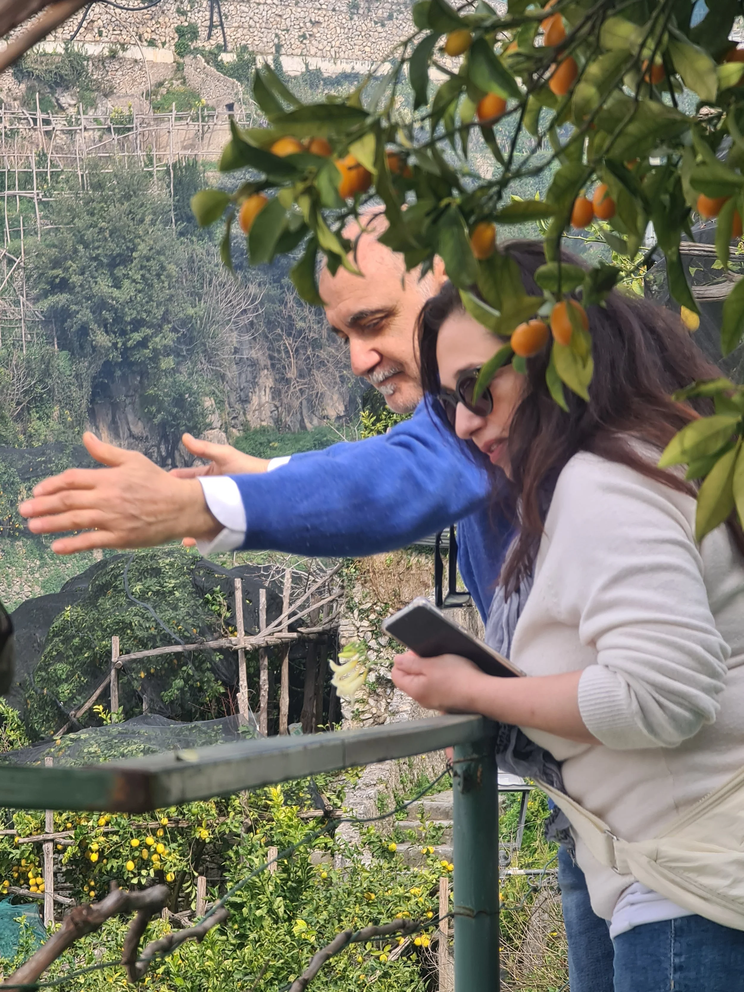 A man and a woman look over a balcony at a scenic outdoor area with trees, yellow fruit, and hills in the background. The man is pointing outward while the woman holds a smartphone.