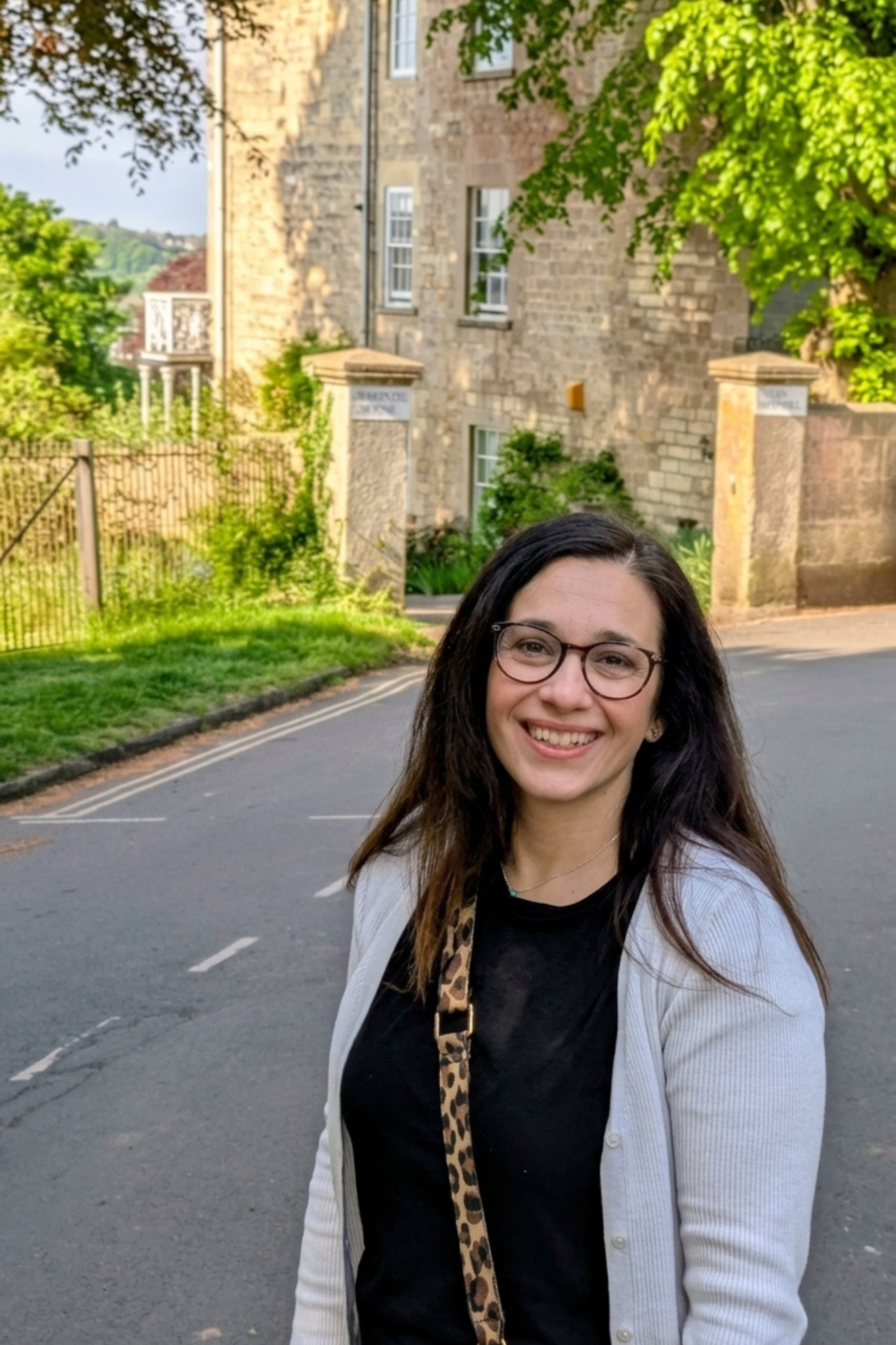 Portrait of a woman with glasses smiling outdoors in front of an old stone building and green trees.
