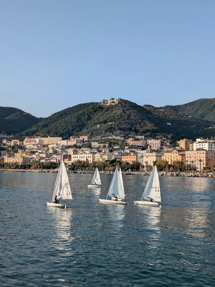 Four small sailboats with white sails sailing on a body of water, with a cityscape and green mountains in the background under a clear blue sky.