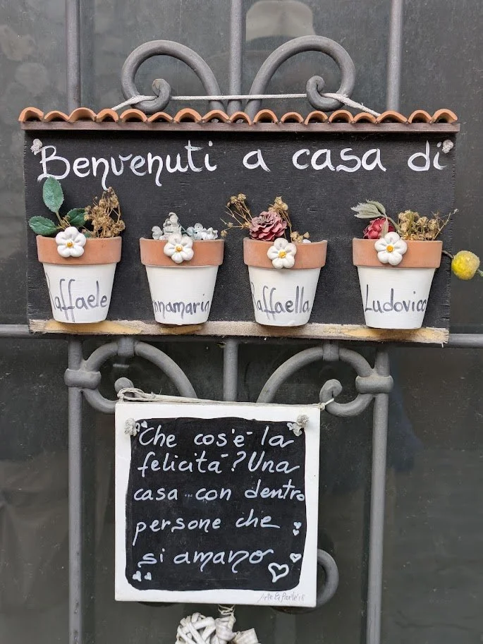 Decorative sign in Italian with four flower pots labeled with people's names and a quote about happiness.