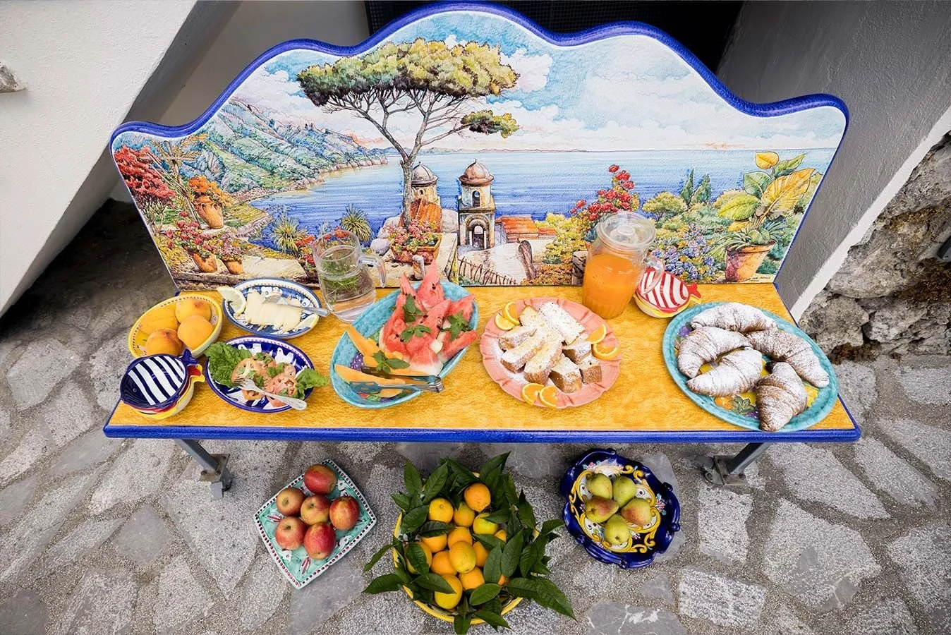 A colorful outdoor breakfast setup on a yellow table with a scenic Mediterranean-style mural backing, featuring fruits like apples and lemons, watermelon slices, croissants, and various breakfast items, with jars of orange juice and water, and decorated with vibrant ceramic dishes and bowls.