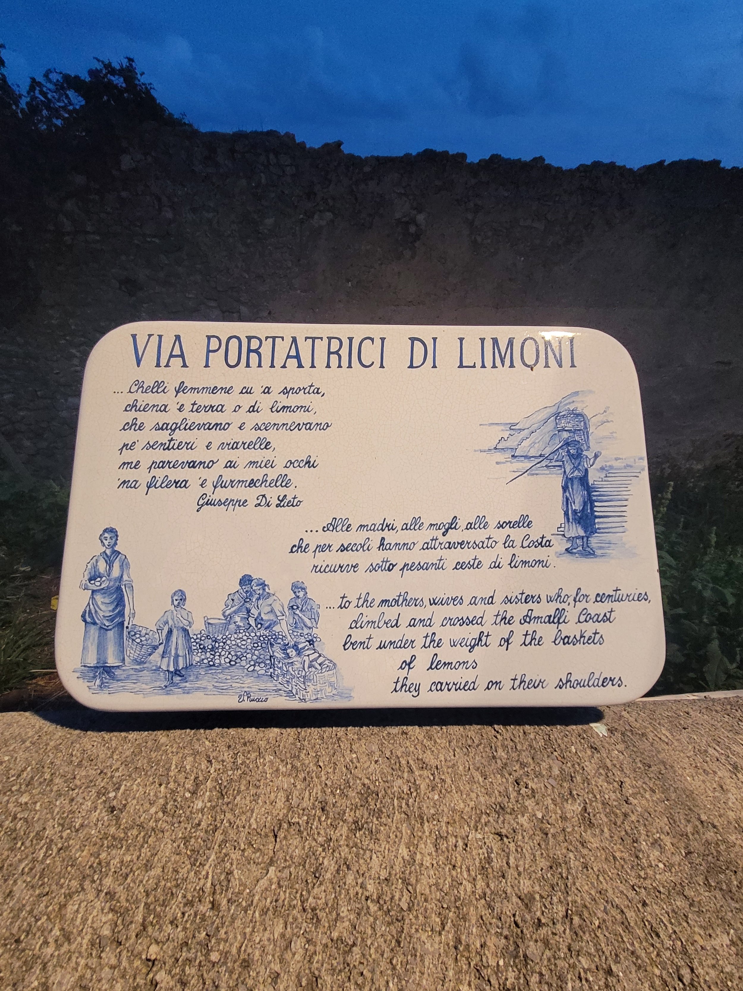 Sign with Italian and English text titled 'Via Portatrici di Limoni', featuring illustrations of women and children carrying baskets of lemons, set outdoors against a natural landscape background.