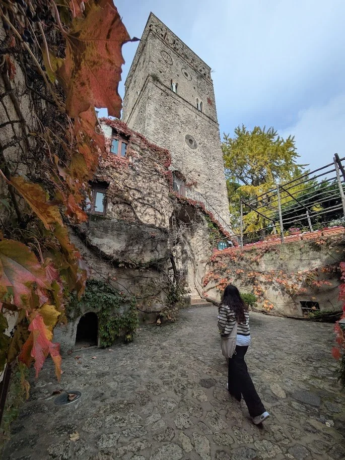 A woman in a striped sweater and black pants walking on a cobblestone path with an old stone tower in the background, surrounded by autumn leaves and foliage.