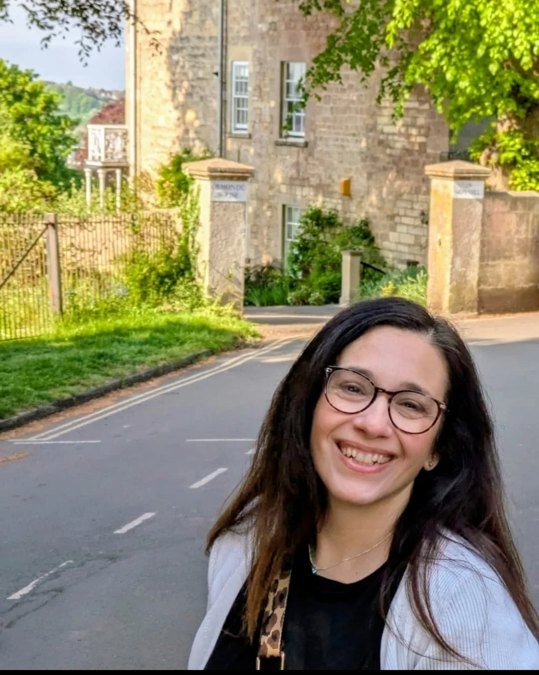A young woman with glasses and long dark hair smiling outdoors in front of a stone building, trees, and a quaint street.