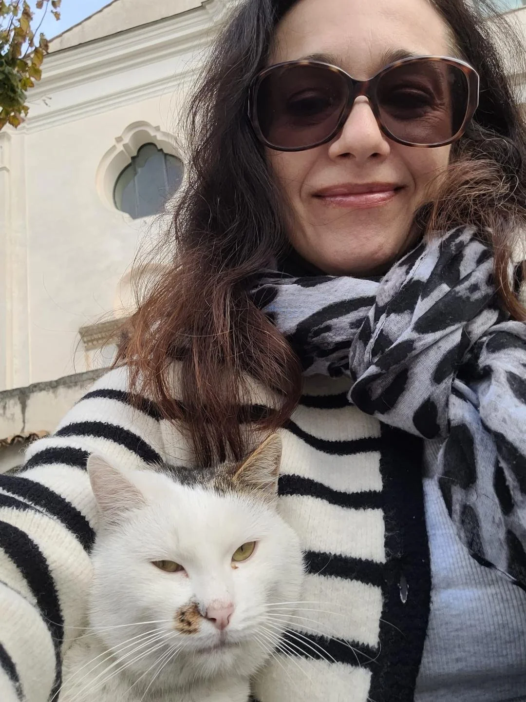 A woman wearing large sunglasses, a striped shirt, and a patterned scarf, smiling, with a white cat with some brown markings resting against her chest, outdoors in front of a white building with a decorative window.