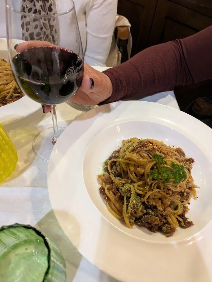 A colorful outdoor breakfast setup on a yellow table with a scenic Mediterranean-style mural backing, featuring fruits like apples and lemons, watermelon slices, croissants, and various breakfast items, with jars of orange juice and water, and decorated with vibrant ceramic dishes and bowls.