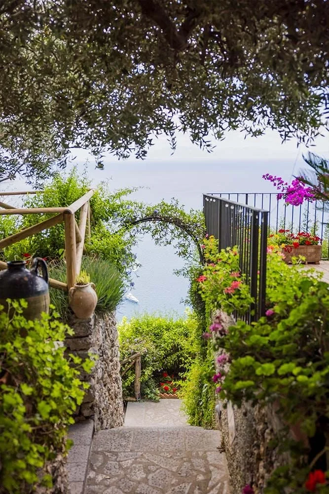 Scene in private villa in Ravello. A stone pathway leads down a garden stairs with lush green plants and colorful flowers on either side. A black railing and an archway covered with vines are visible, with a view of the sea and sky in the background.