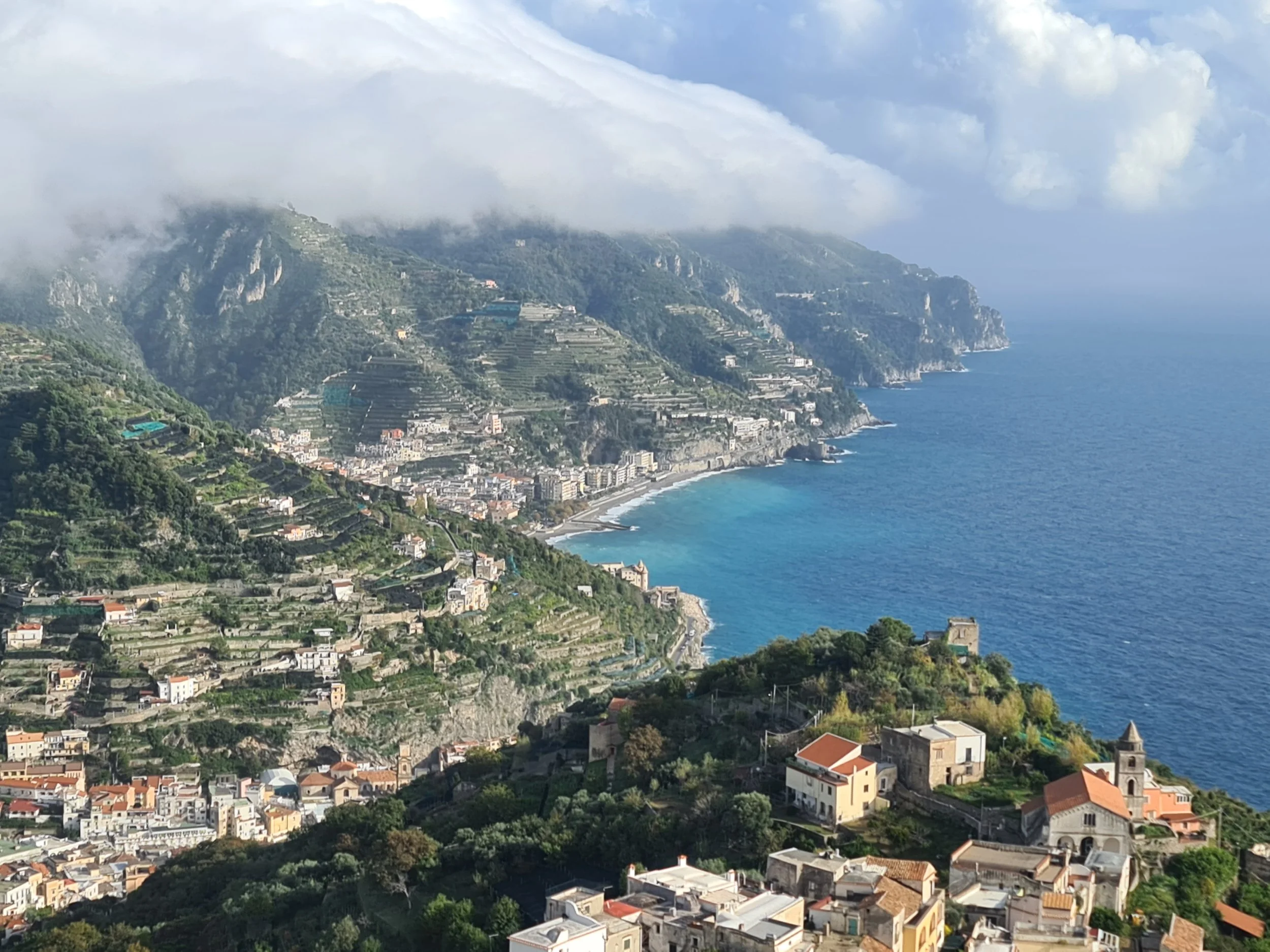 The Amalfi Coast from Ravello nestled on green hills with terraced farmland, a church with a bell tower, and a view of the sea with waves and rocky cliffs, under a partly cloudy sky.