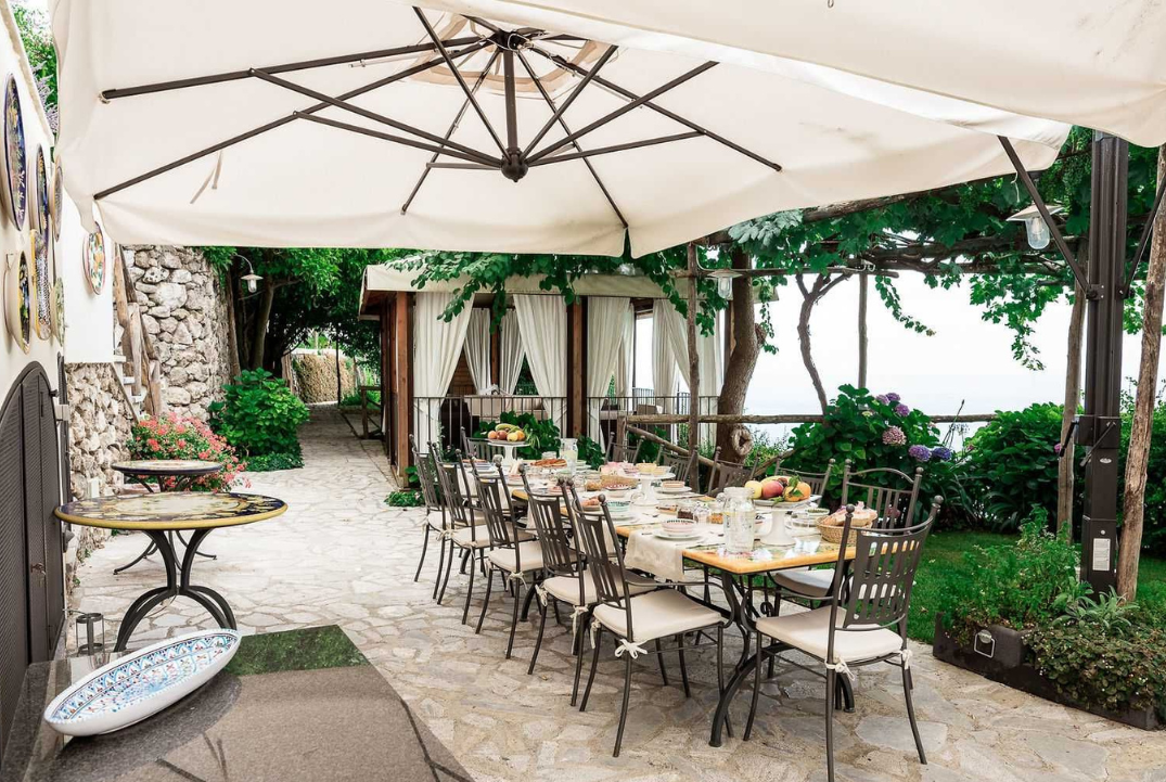 An outdoor dining setup with a long table covered with a white tablecloth, set for a meal with dishes, glasses, and fruit. Large umbrellas provide shade, and there are cozy cabana-style structures draped with white curtains in the background. The area is surrounded by lush greenery and flowering plants.